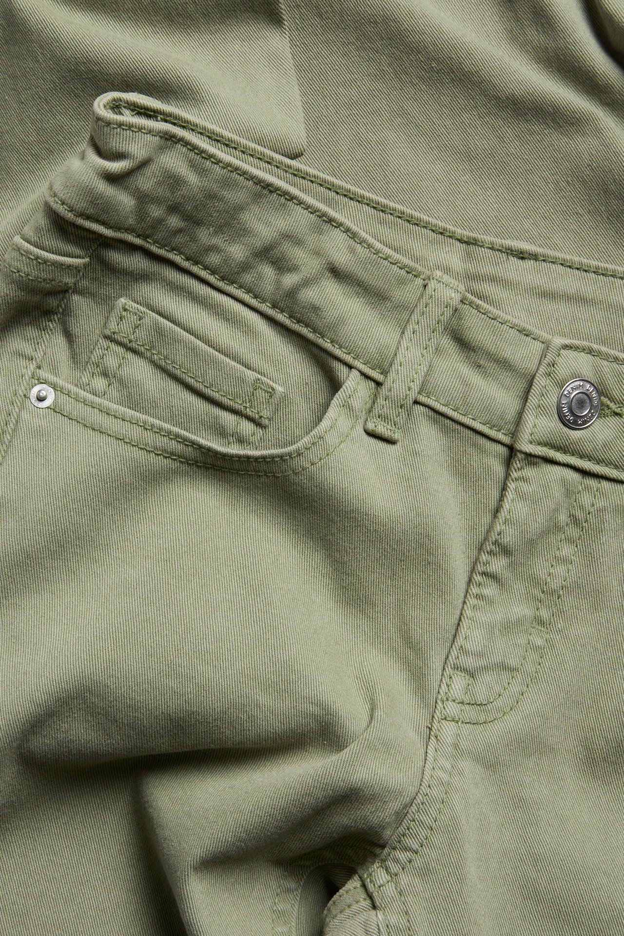 Nærbilde av mørkegrønne bootcut-jeans med lav midje, som viser frontlommen og en metallknapp.