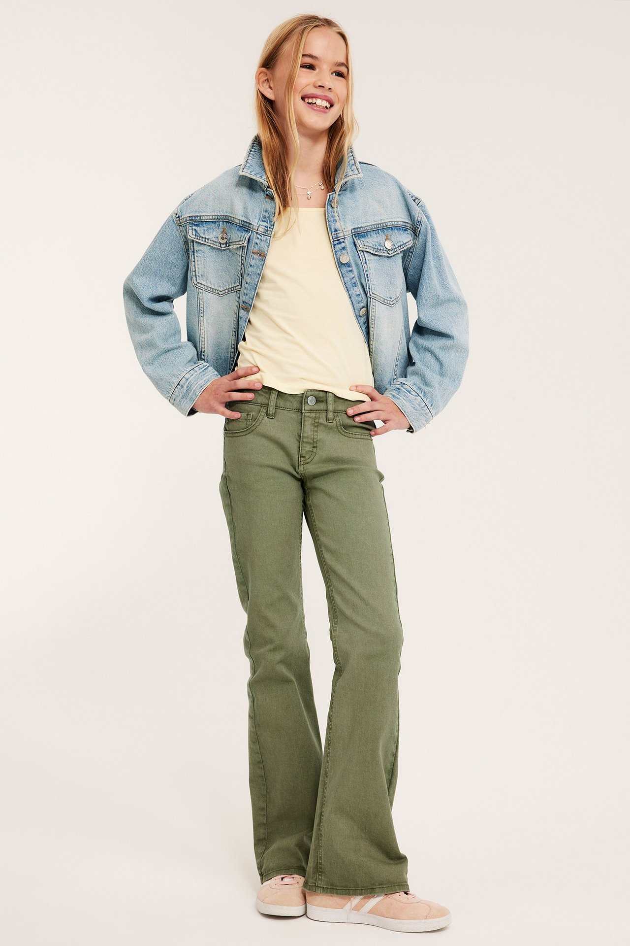 Edestä: lapsi tummanvihreissä matalavyötäröisissä bootcut-farkuissa, vaaleankeltaisessa topissa ja vaaleansinisessä farkkutakissa.