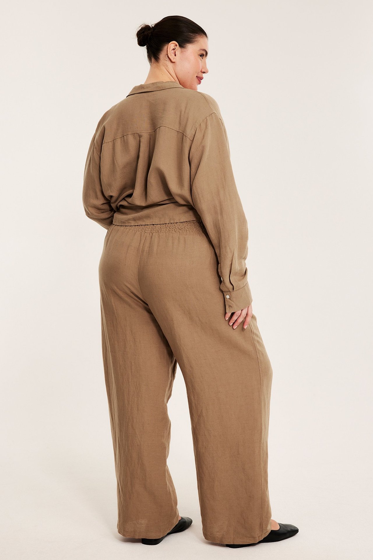 Ryggvy: kvinna i beiga vida byxor i linneblandning med smockresår i midjan och matchande långärmad skjorta.