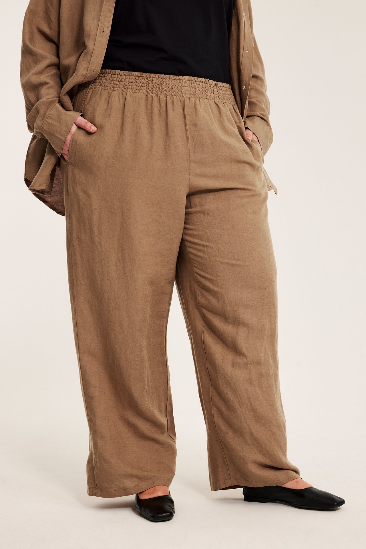 Framifrån: kvinna i beiga vida linnebyxor med smockresår i midjan, öppen beige skjorta och svarta platta skor.