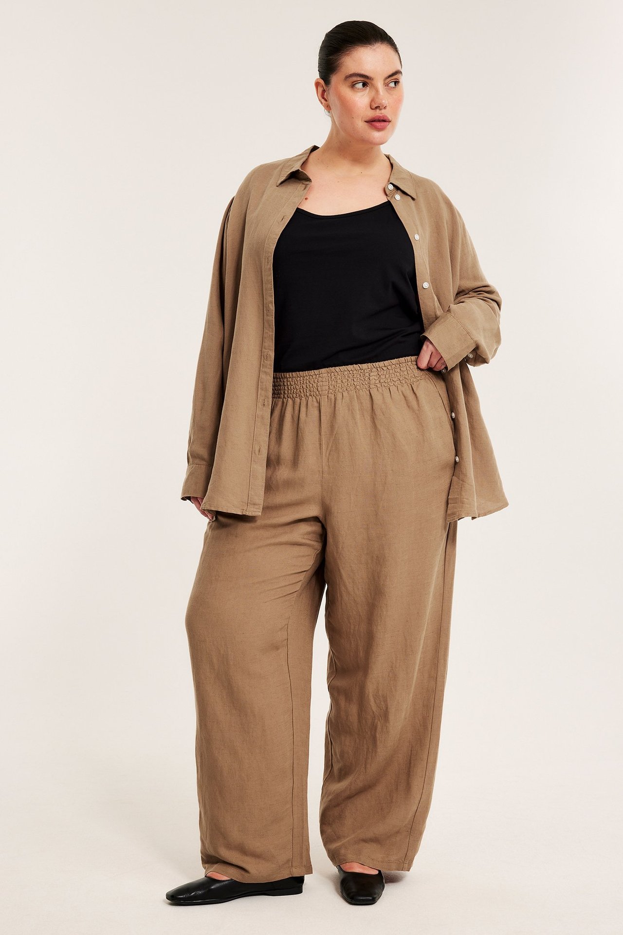 Beiga pull-on linnebyxor med smockmidja, bärs av kvinna med öppen beige skjorta och svart topp.