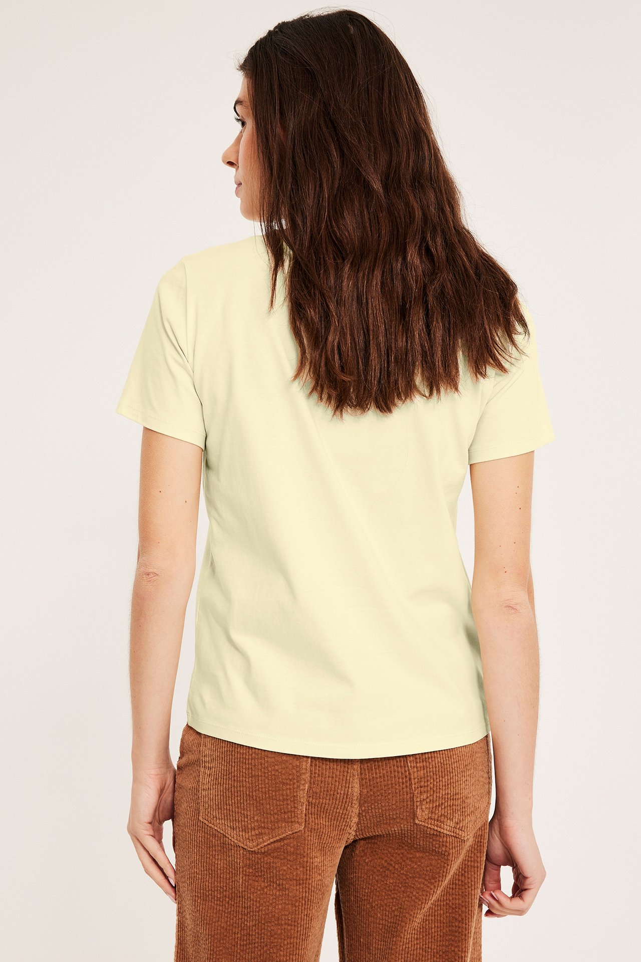 Naisen takakuva: vaaleankeltainen, suora puuvilla-T-paita ja ruskeat vakosamettihousut.