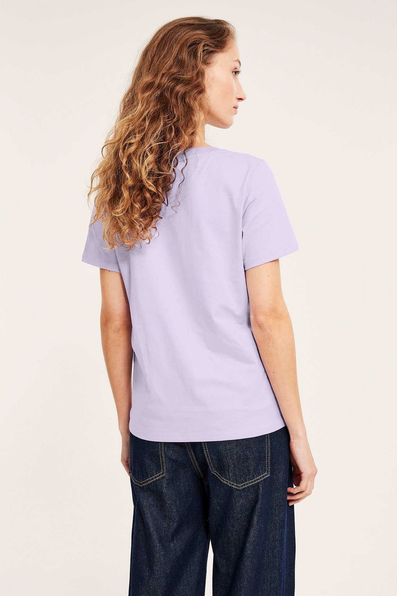 Naisen selkäpuolen näkymä yllään vaaleanliila suoraryhtinen puuvillainen t-paita ja tummansiniset farkut.