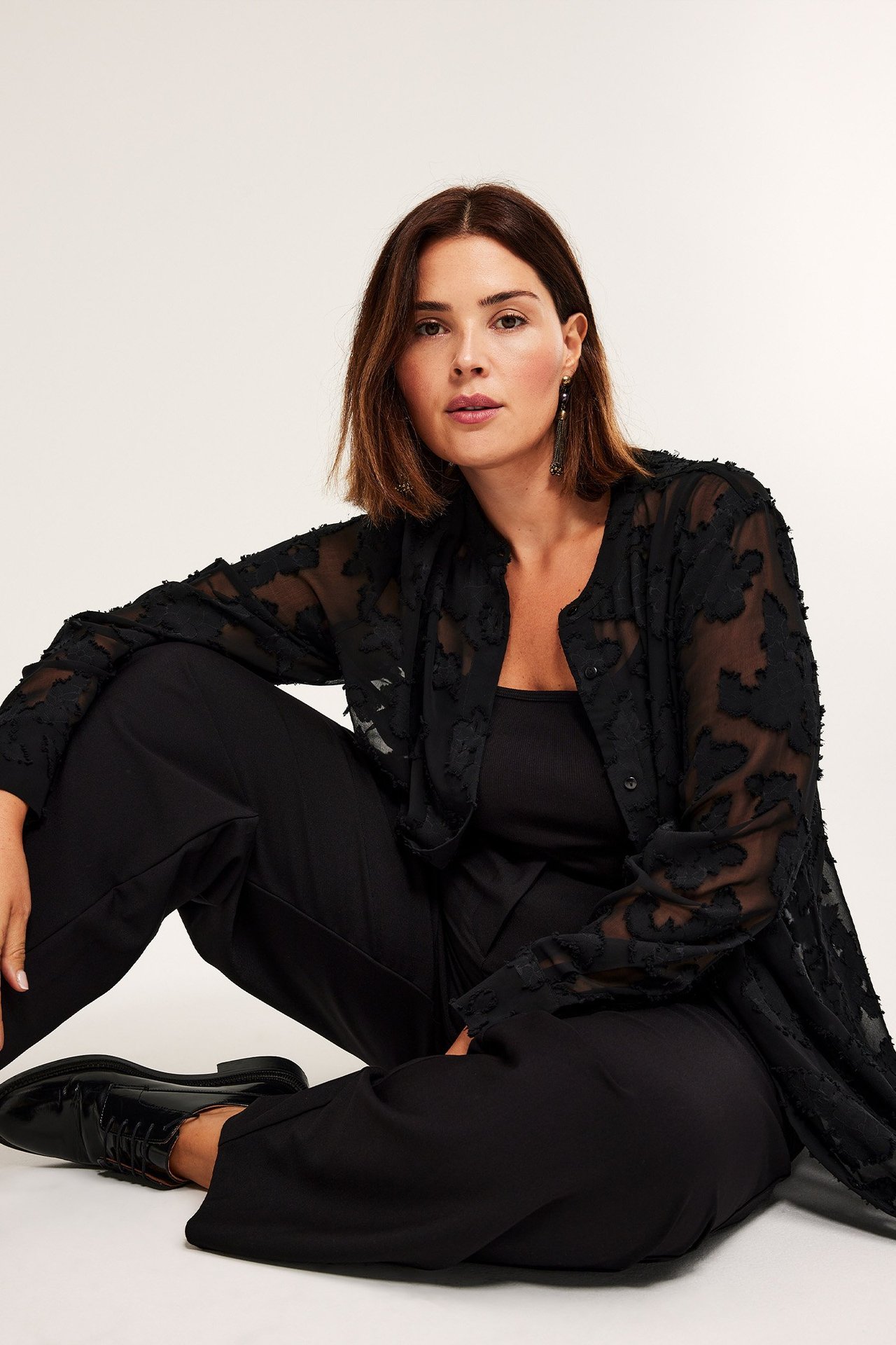 Nainen edestä avoimessa mustassa läpikuultavassa jakardipaidassa, topissa, mustissa housuissa ja nauhakengissä.