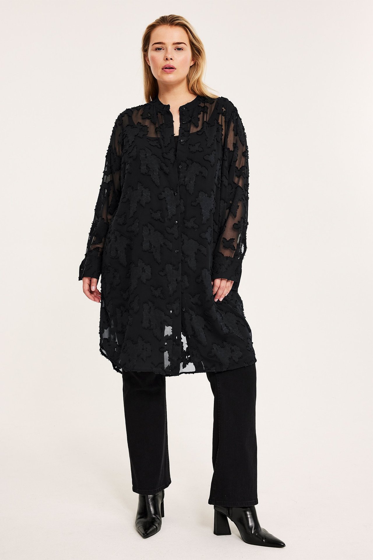 Frontvy: kvinna i svart jacquard långskjorta, svarta byxor och svarta ankelboots.