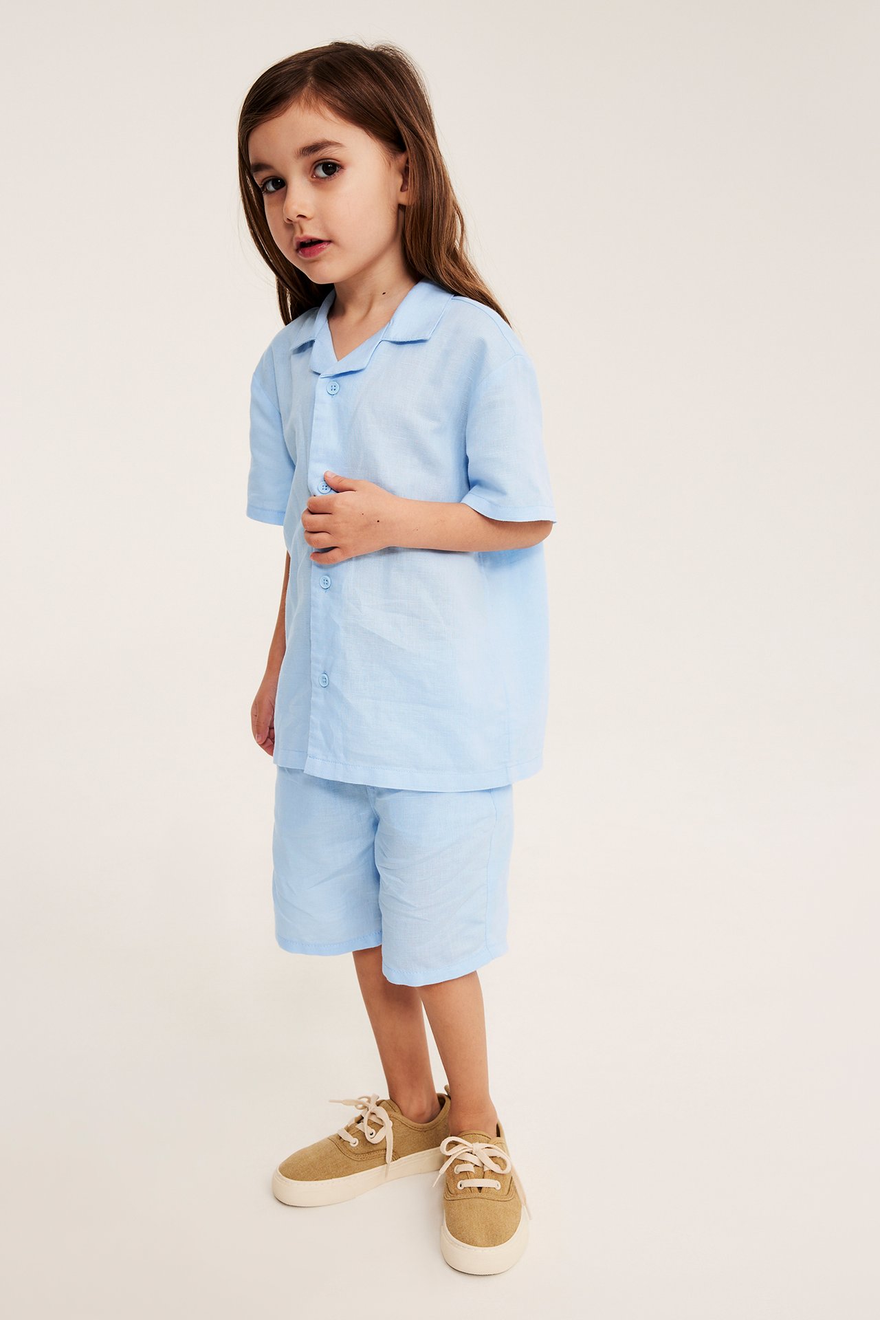 Widok z przodu dziecka w jasnoniebieskich szortach z lnu, pasującej koszuli i beżowych sneakersach.