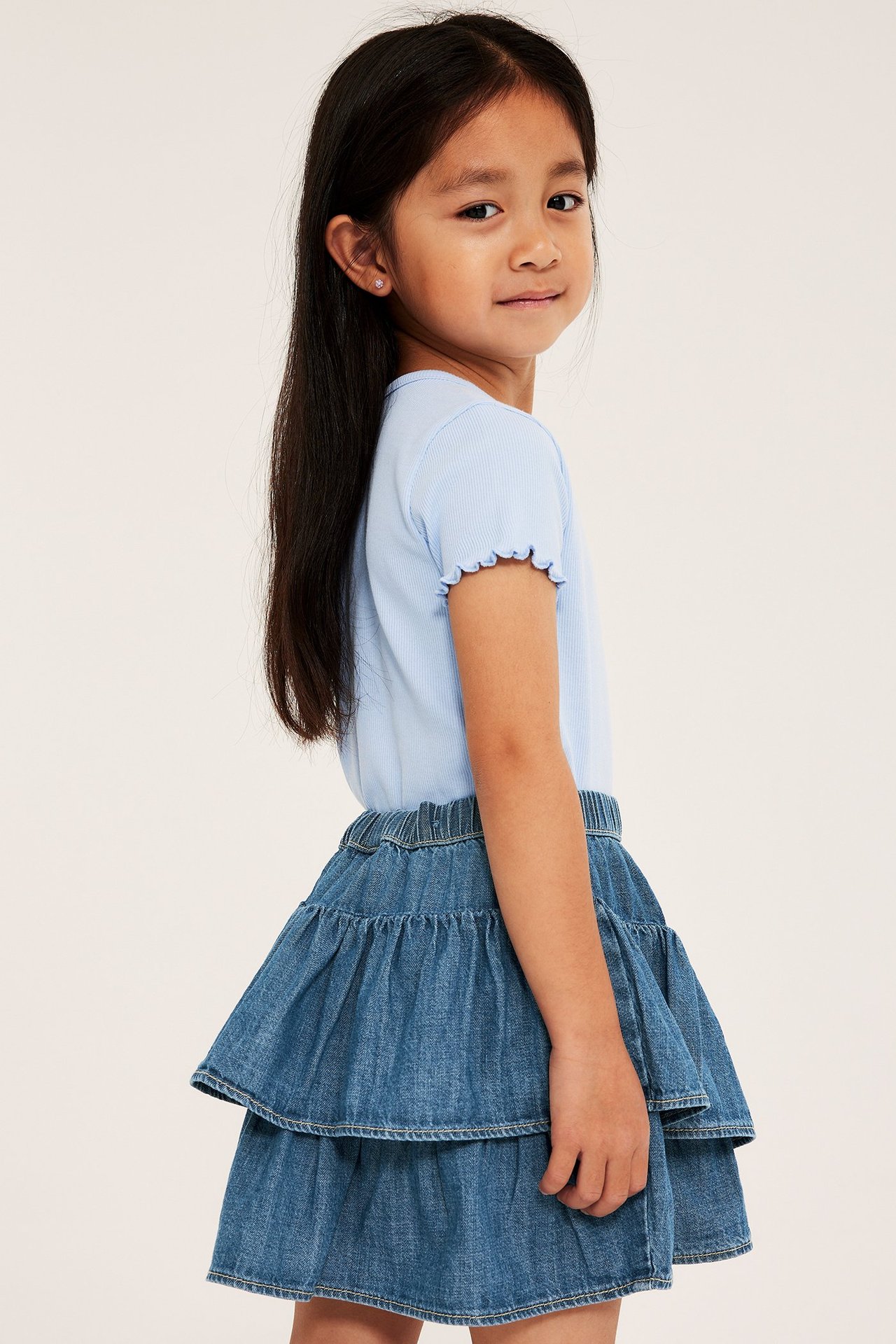 Sivukuva lapsesta vaaleansinisessä kerroksellisessa farkku-röyhelöhameessa ja vaaleansinisessä ribbipaidassa.