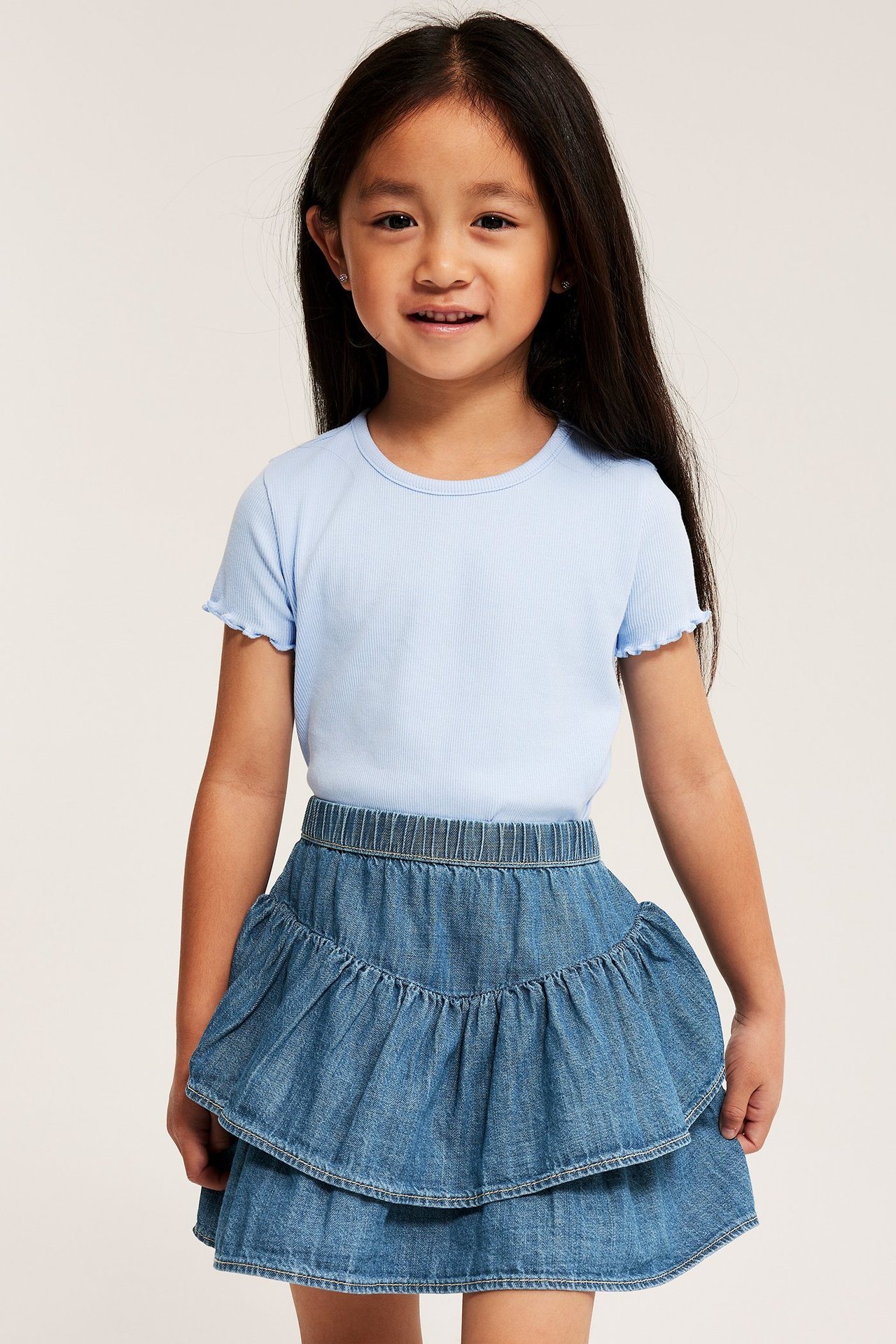 Front view: Ett barn i en ljus denim volangkjol och en ljusblå ribbad t-shirt.