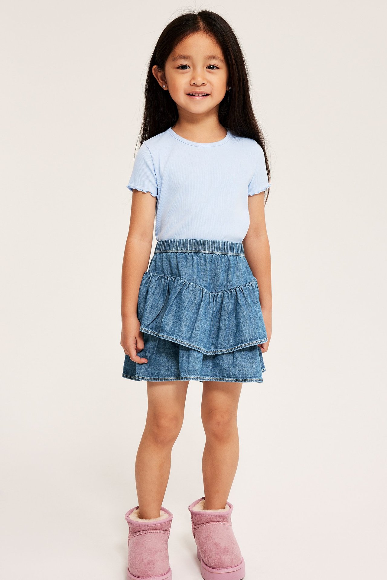 Front view: Kid i ljus denim volangkjol med resårmidja, ljusblå t-shirt och rosa stövlar.