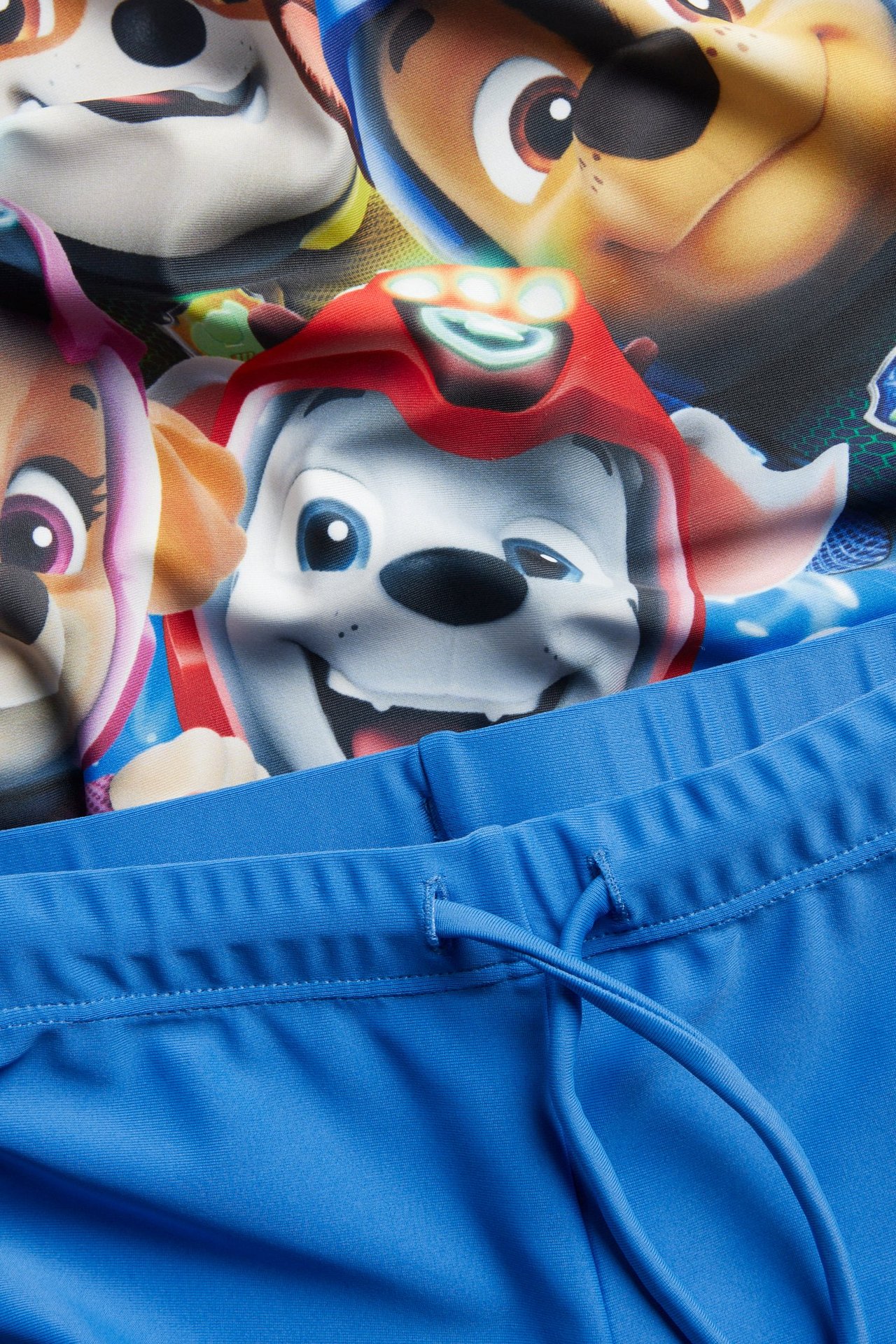 Lähikuva, lasten UV-setti: monivärinen Ryhmä Hau -printtitoppi ja yksiväriset siniset uimahousut kiristysnyörillä.