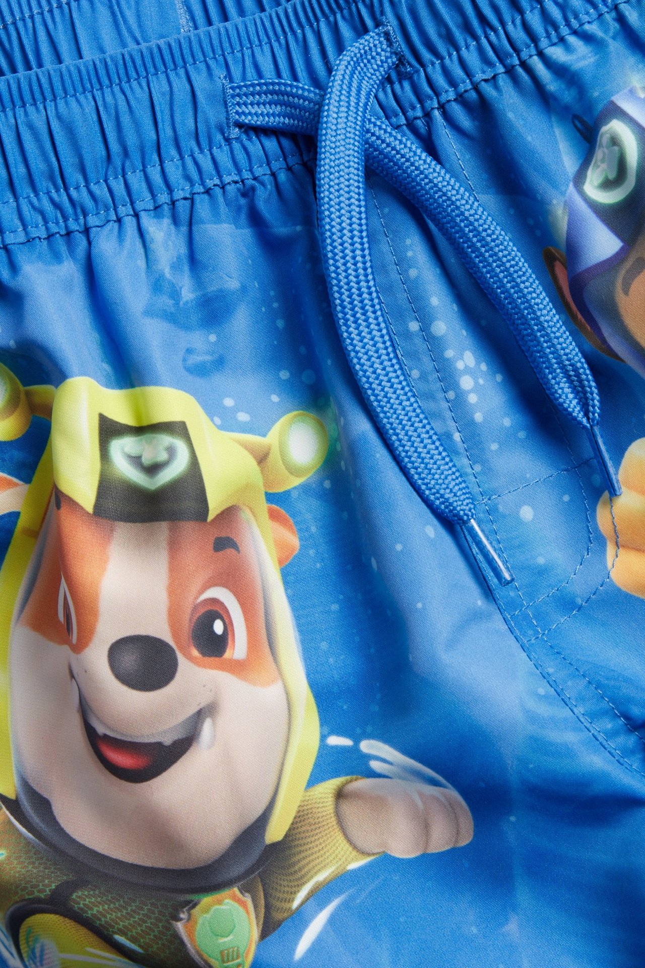 Nærbilde, forfra, av blå badebukser for barn med Paw Patrol-trykk og elastisk snøring i midjen.