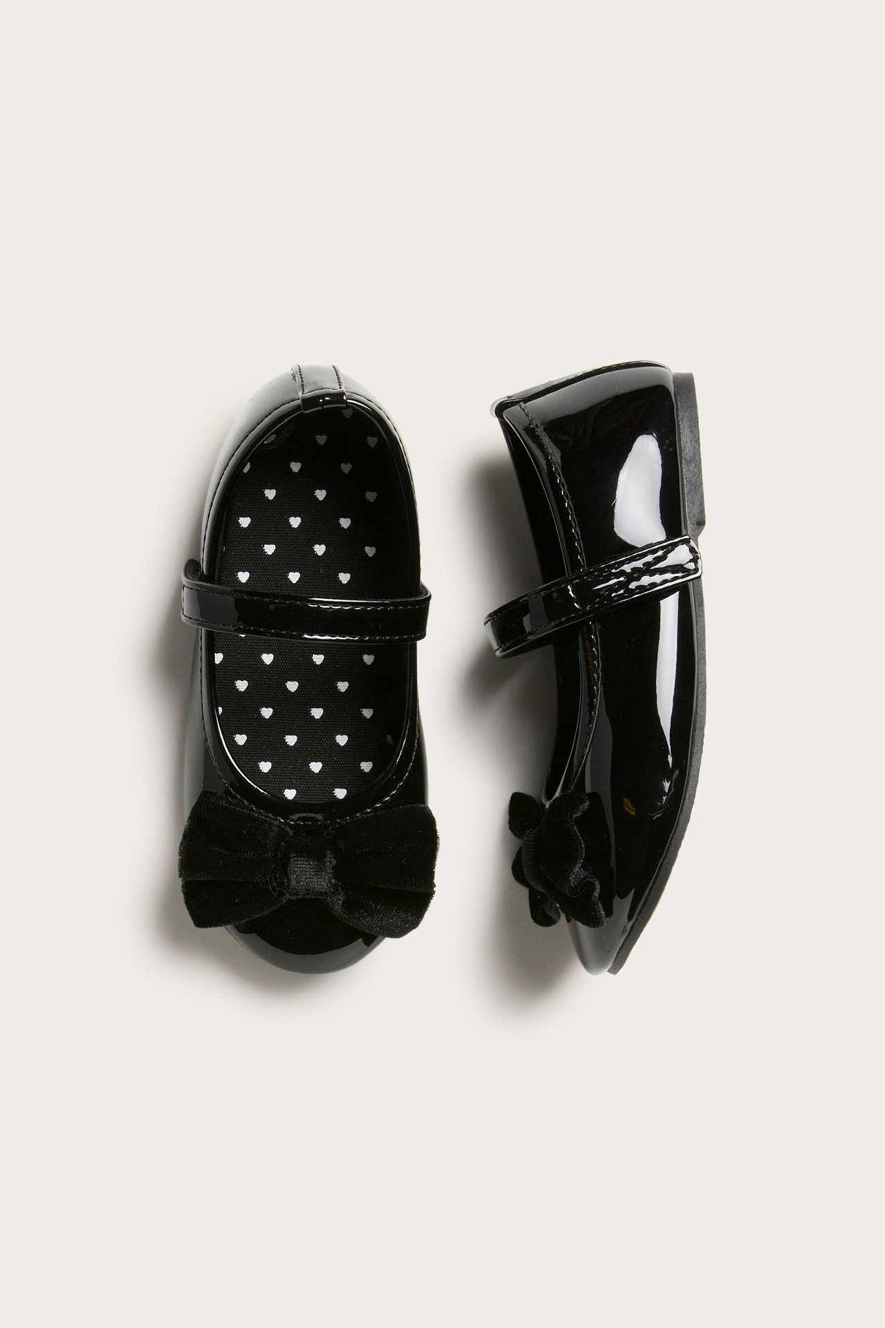 Widok z góry: dwie czarne lakierowane balerinki dziecięce z aksamitnymi kokardkami i serduszkowymi wkładkami.