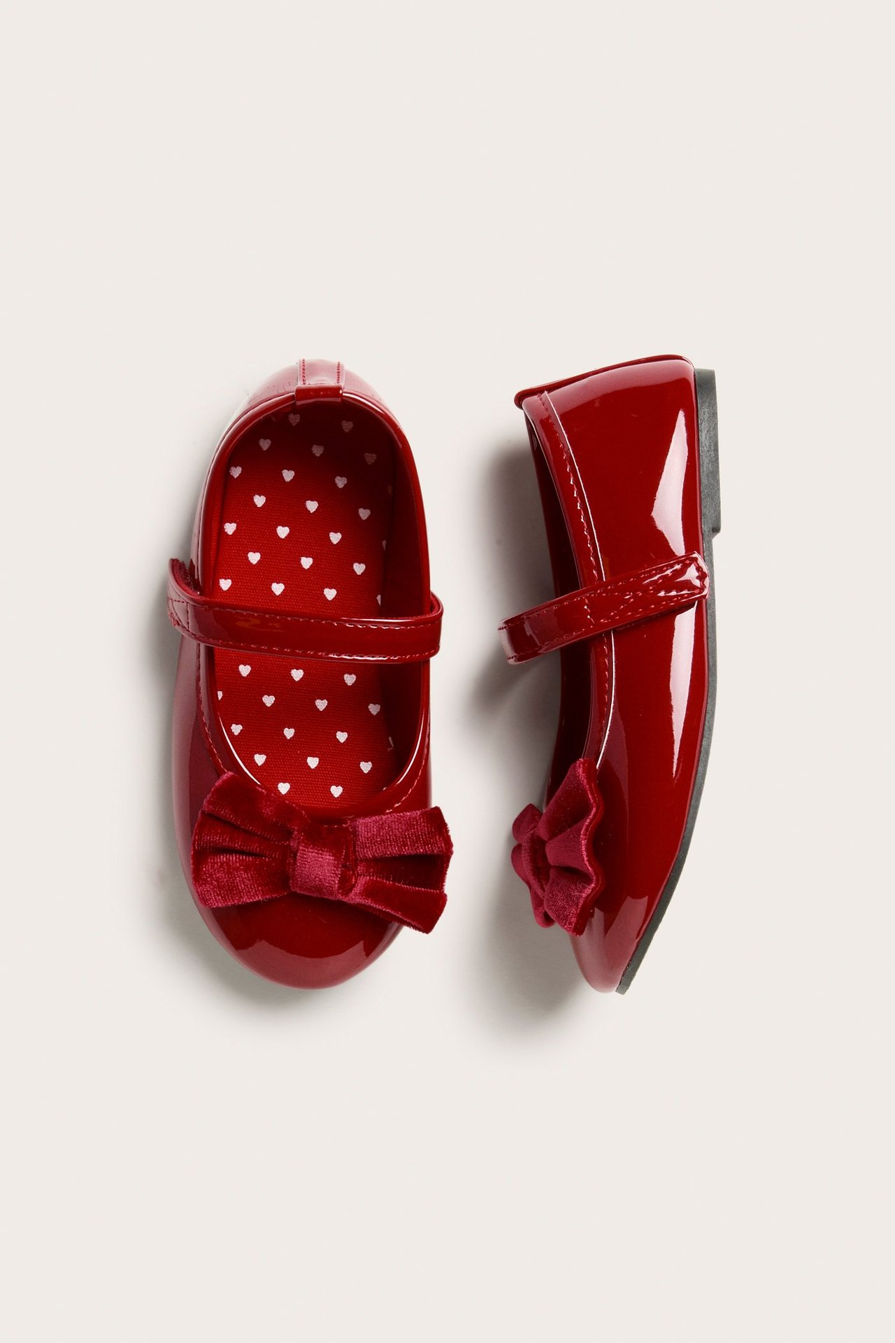 Ylhäältä kuvattuna kaksi punaista kiiltonahkaista ballerinakenkää remmeillä, samettiruseteilla ja sydänkuvioiduilla pohjallisilla.
