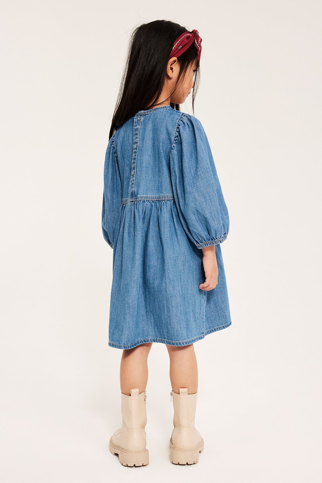 Widok z tyłu: dziecko w jasnoniebieskiej sukience jeansowej z bufiastymi rękawami, czerwonej opasce i beżowych butach.