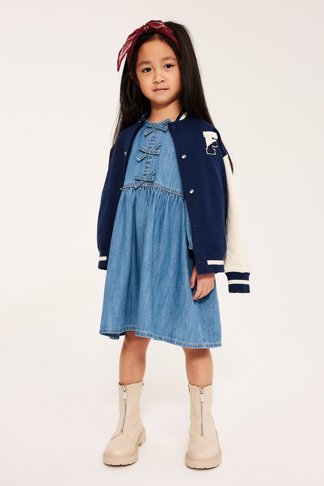 Kid bär en ljus jeansklänning med rosetter, marinblå collegejacka, röd bandana och beiga kängor.