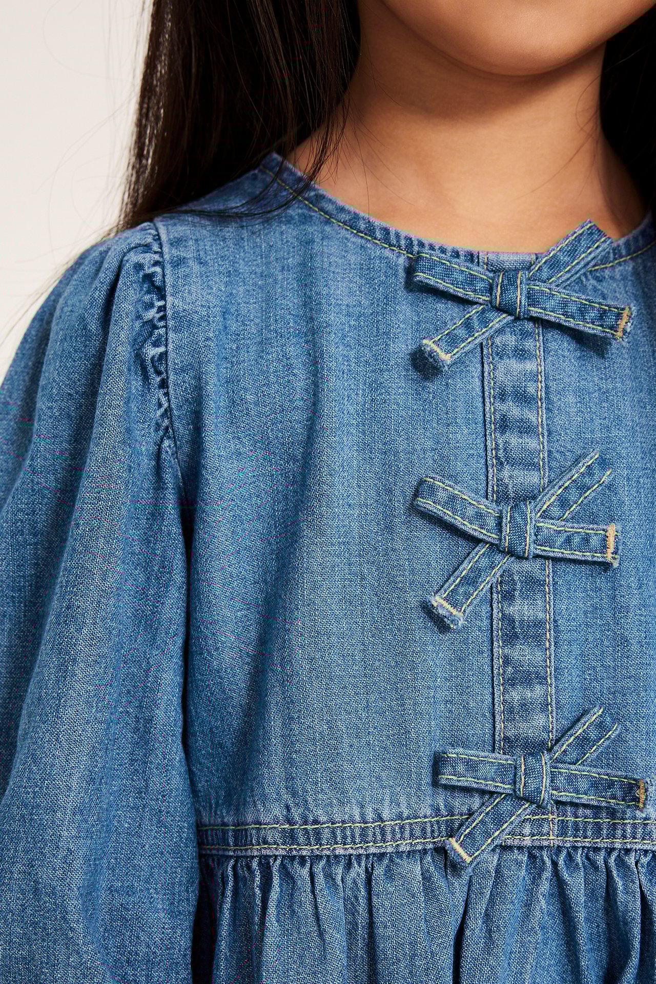 Z bliska, przód: Dziecko w jasnej jeansowej sukience z bufiastymi rękawami i trzema małymi dżinsowymi kokardkami.