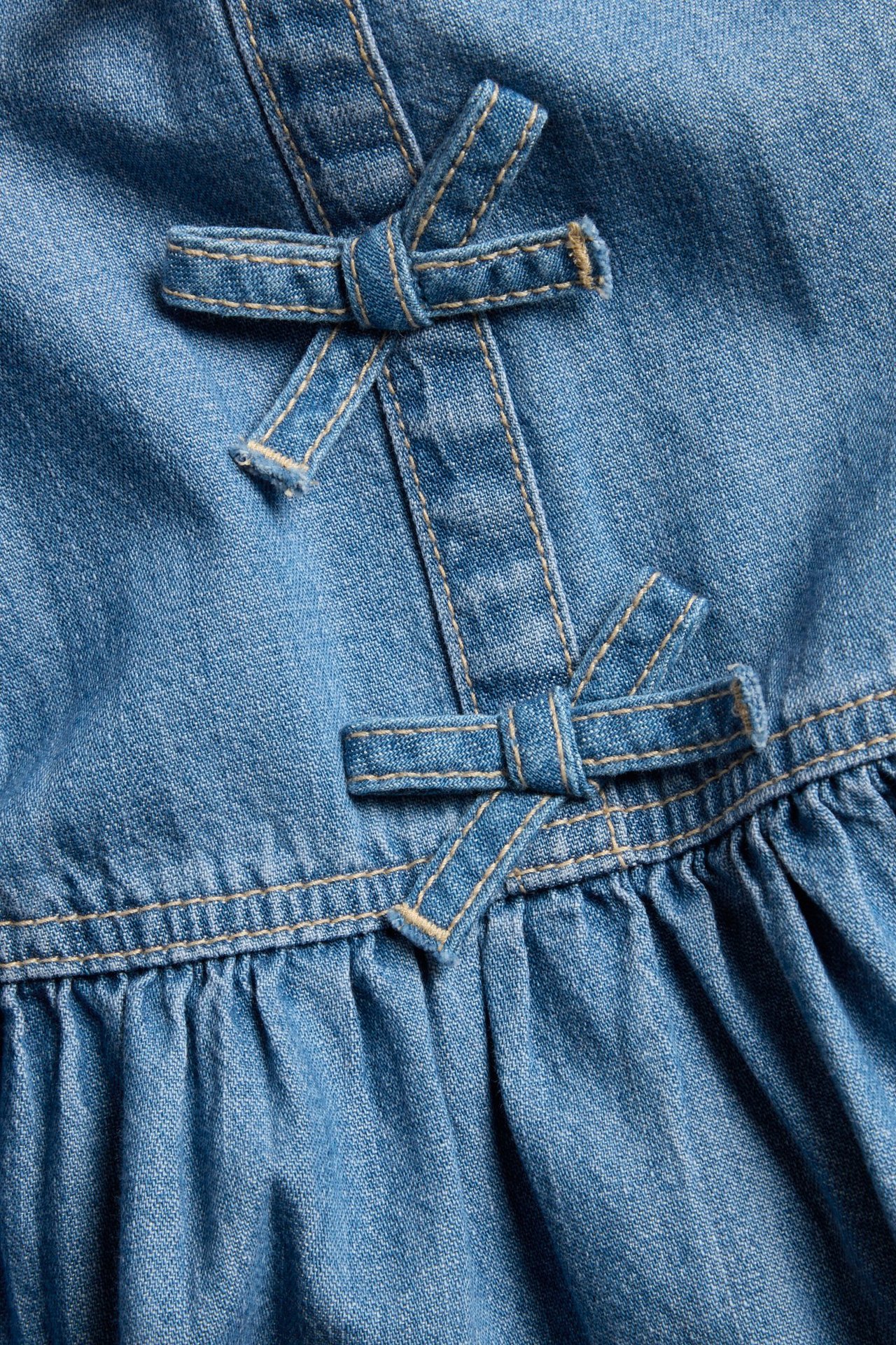 Zbliżenie na jasną jeansową sukienkę z dwiema ozdobnymi kokardami z przodu i marszczonym detalem.