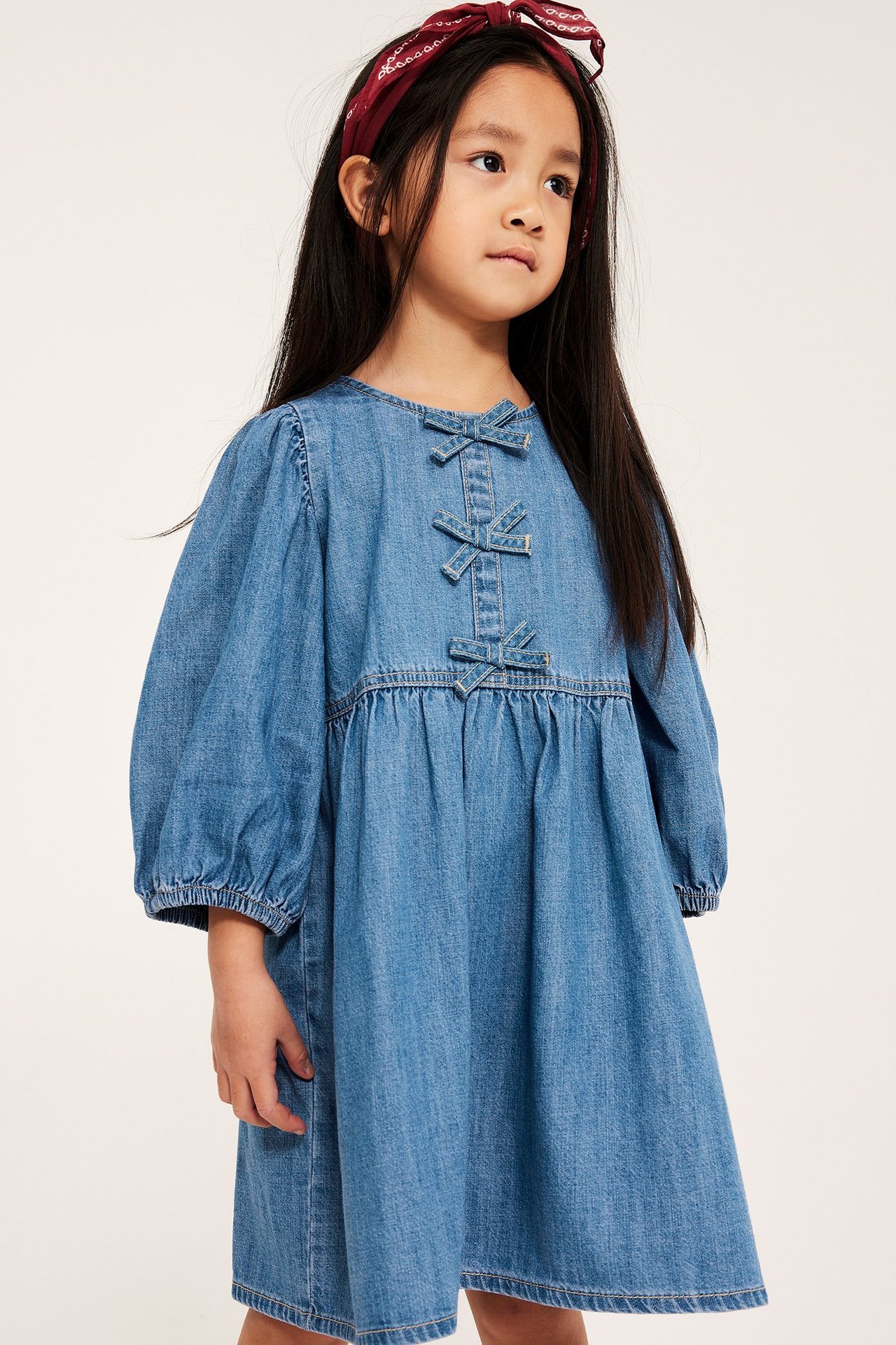 Framsida: Barn i ljus jeansklänning med tre rosetter fram, puffärmar och rött mönstrat hårband.