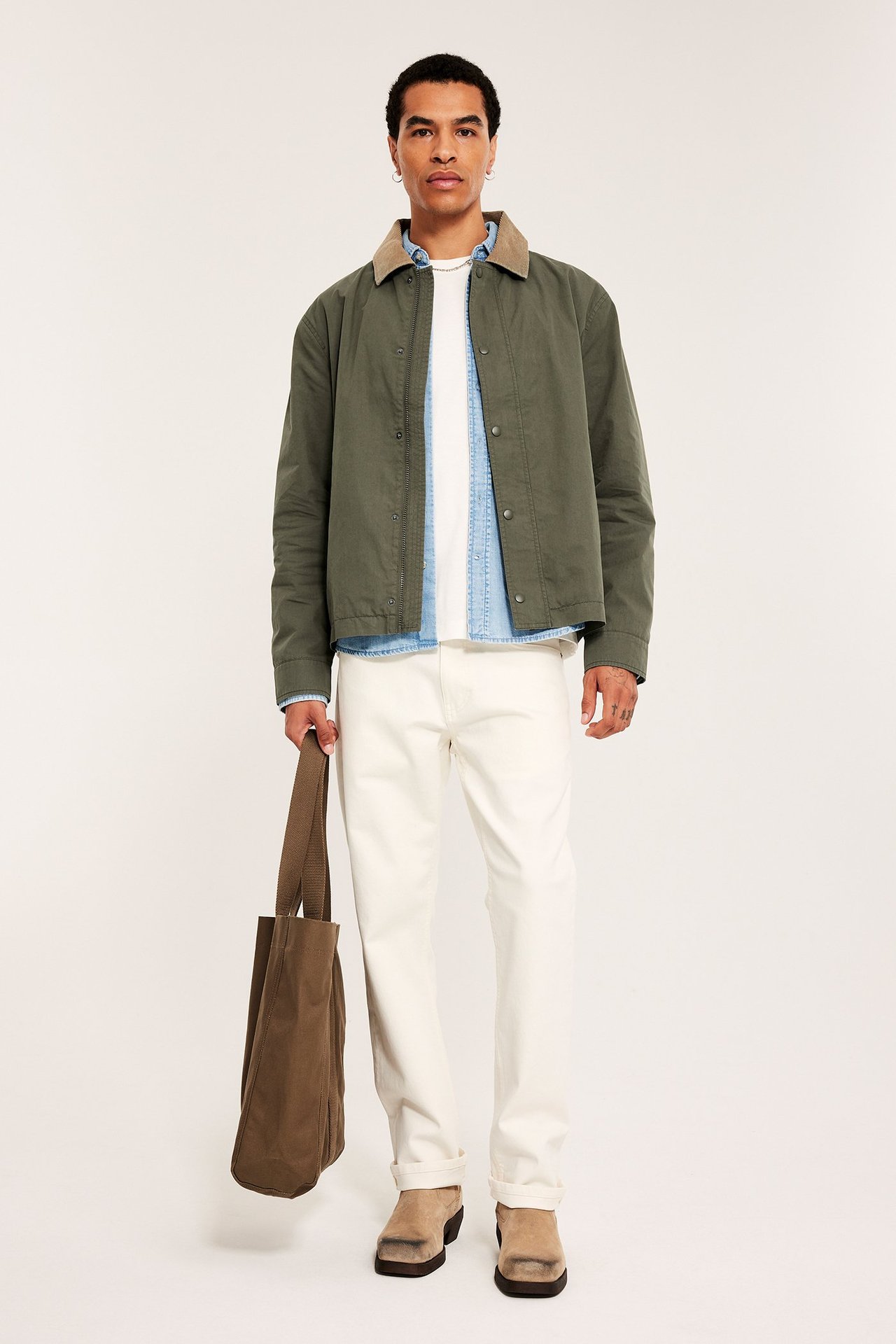 Edestä: Mies pukeutuu vihreään takkiin samettikauluksella, vaaleansiniseen paitaan, valkoisiin housuihin, pitäen ruskeaa kangaskassia.