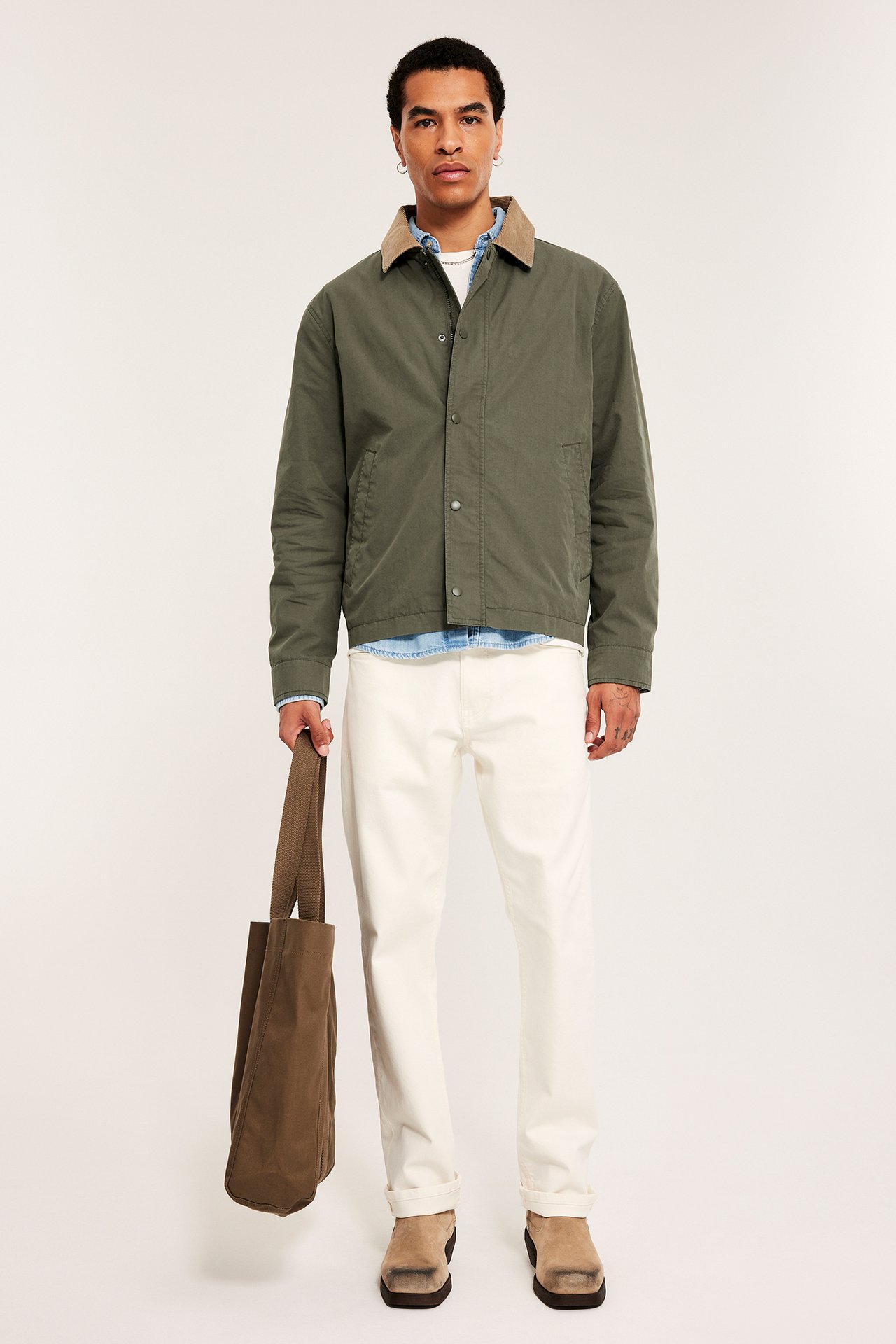 Mężczyzna w zielonej kurtce z kołnierzem sztruksowym i guzikami, kremowych spodniach, beżowych mulach, trzyma brązową torbę.