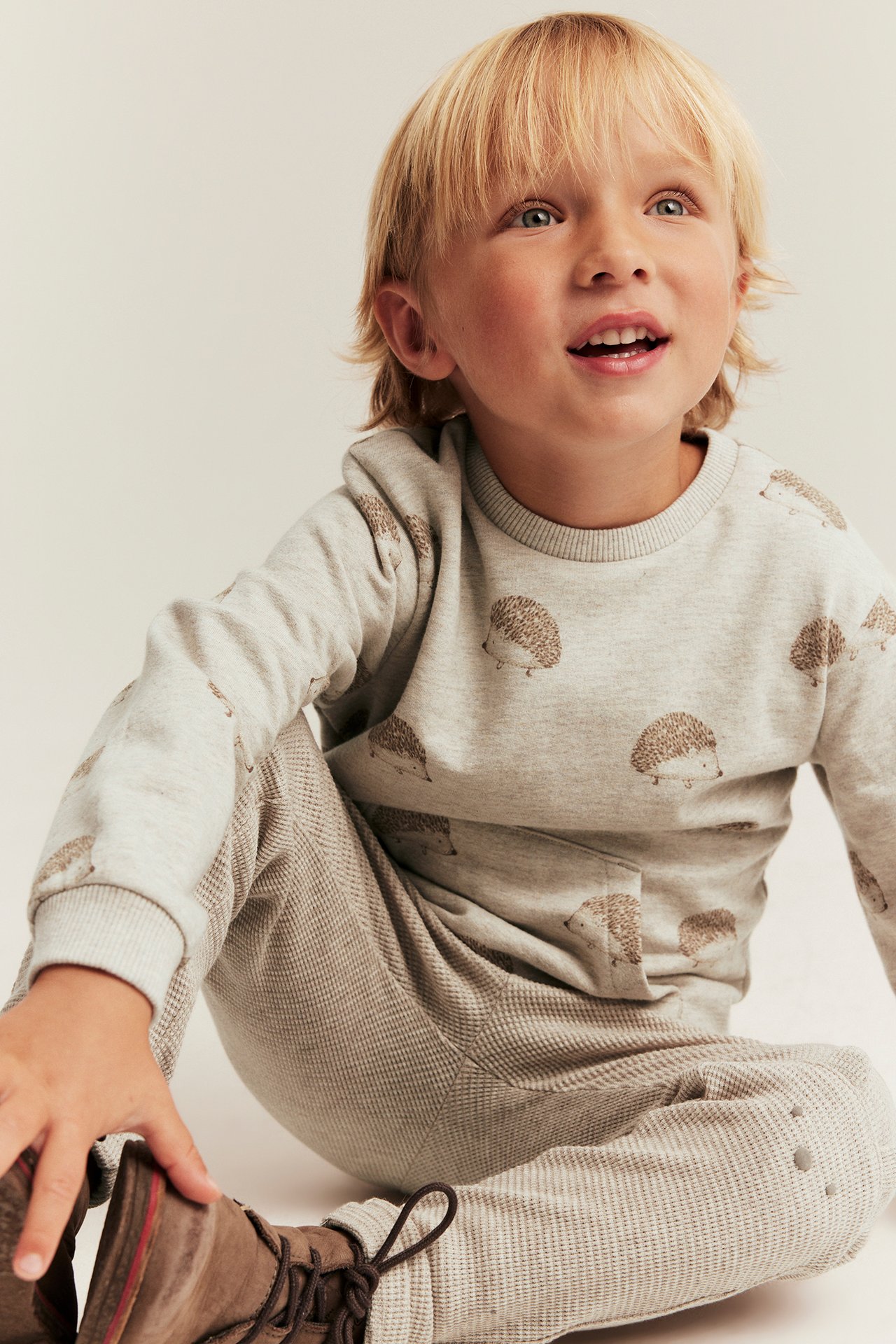 Front view av ett barn i en beige igelkottsmönstrad sweatshirt, beiga byxor och bruna kängor.