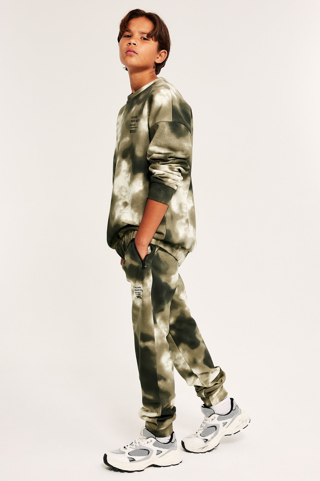 Widok z boku: dziecko w zielonej bluzie dresowej we wzory, spodniach i białych sneakersach.