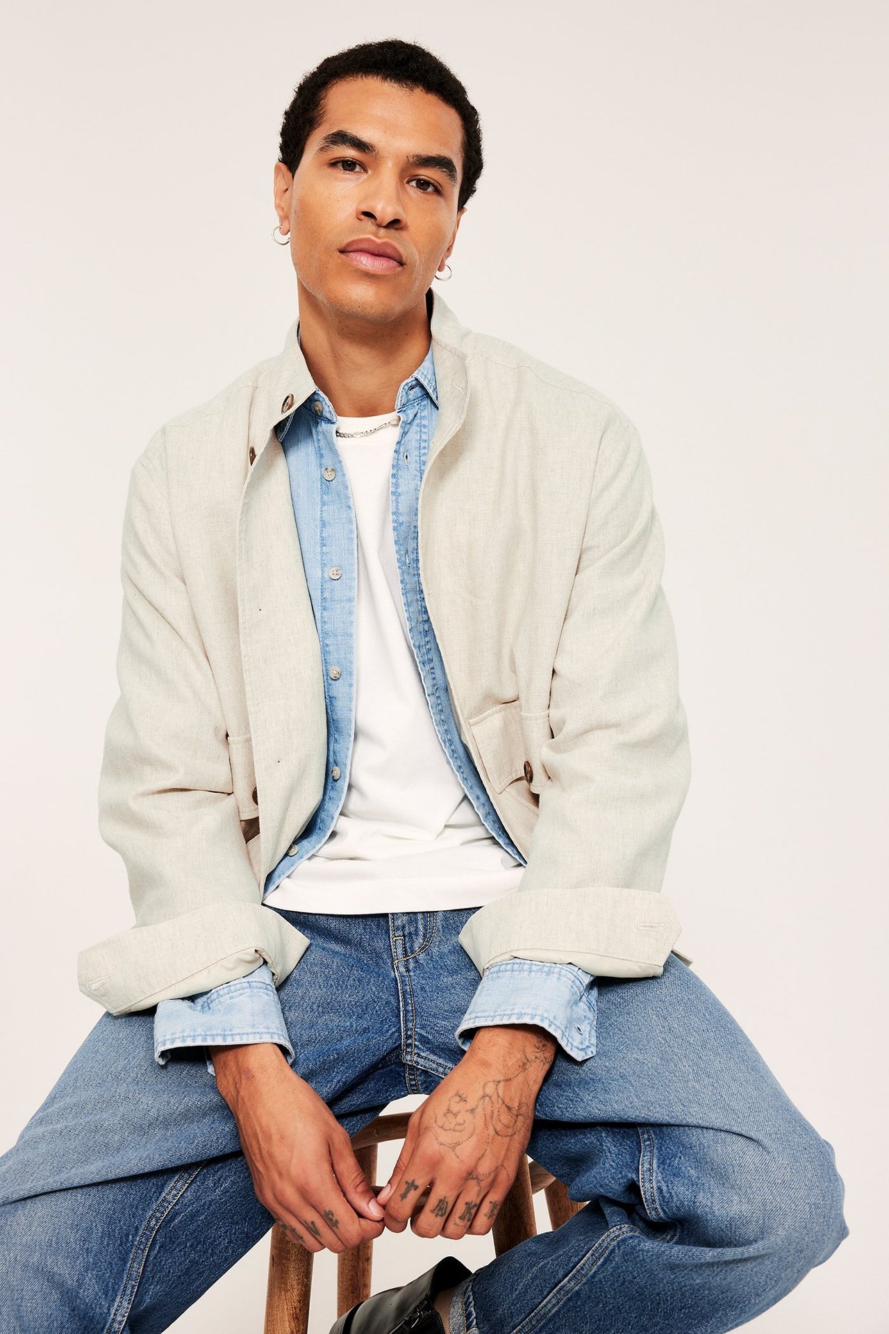 Mies yllään vaaleanbeigessä pellavasekoitetakissa, sinisen farkkupaidan, valkoisen t-paidan ja sinisten farkkujen päällä.