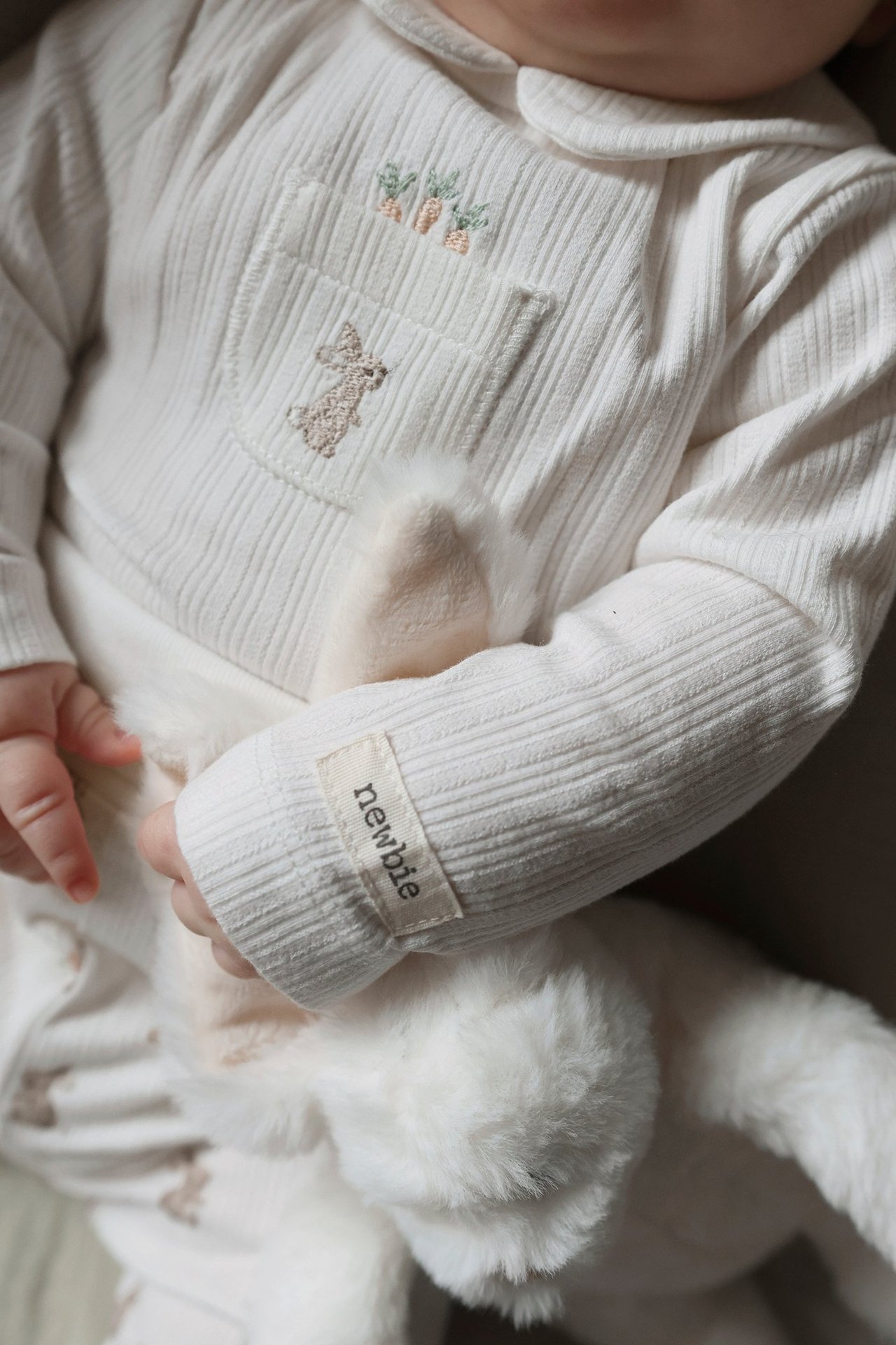Nærbilde av baby i offwhite ribbet langermet body med brodert lomme, holder en hvit kaninleke.