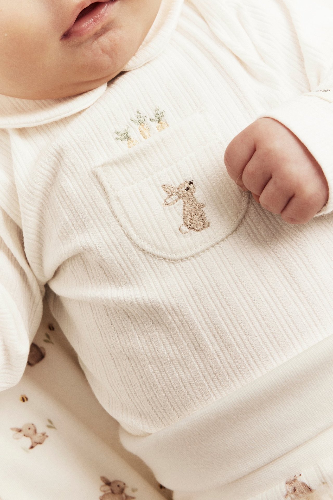 Nærbilde av baby i offwhite ribbet langermet body med krage og brodert kaninlomme.