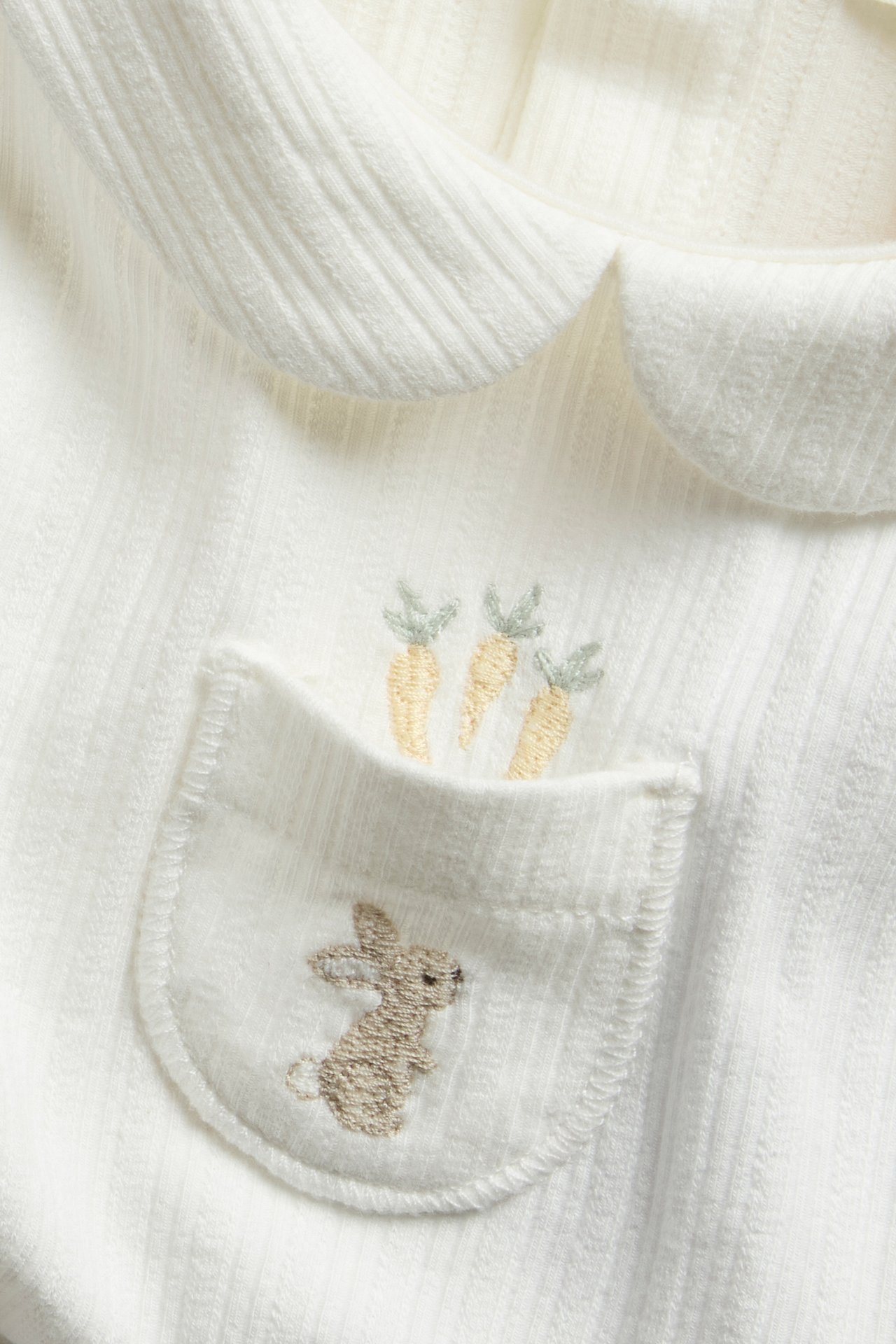 Nærbilde av offwhite ribbet langermet baby-body med krage og brodert kaninlomme.