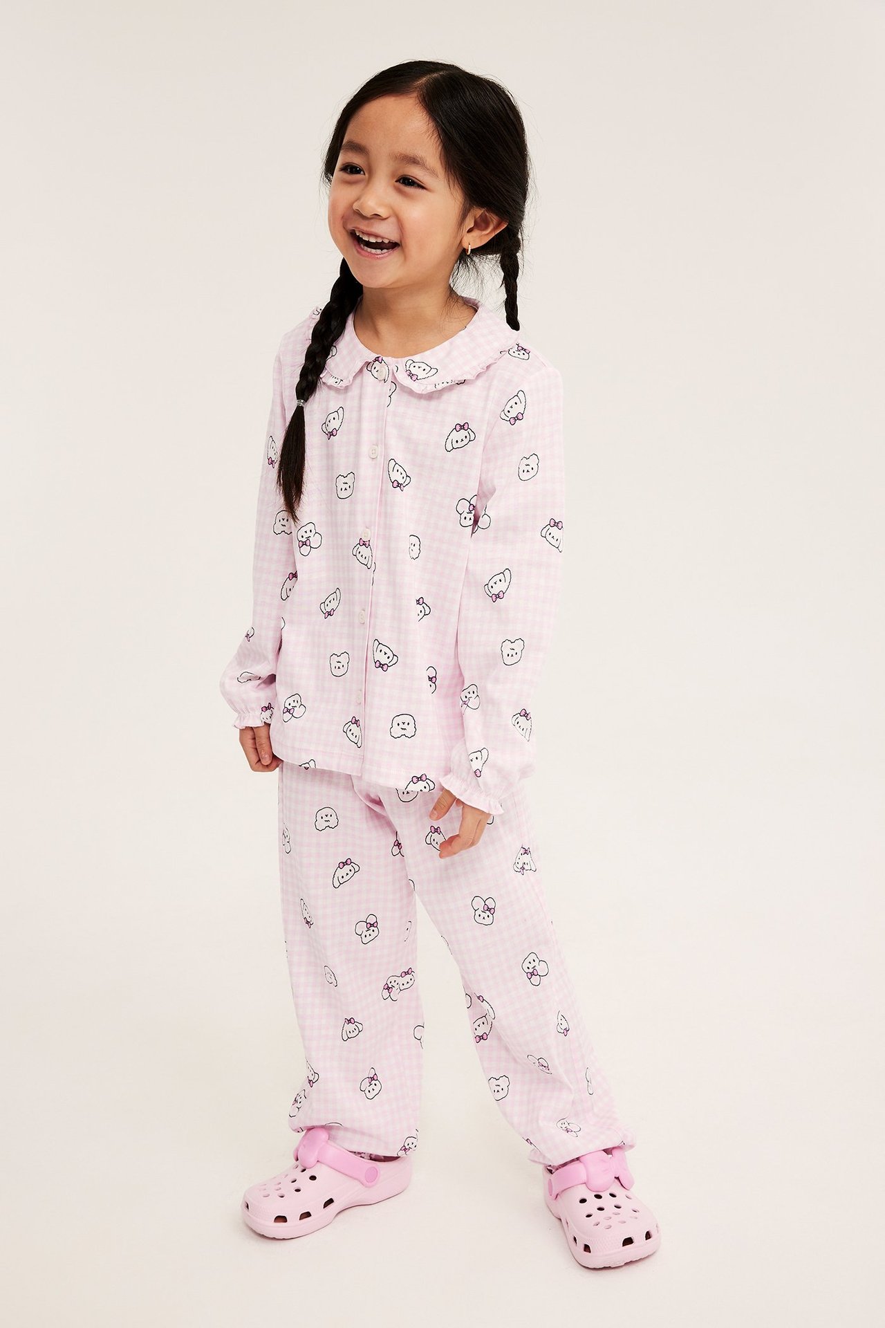 Edestä: Tyttö vaaleanpunaisessa kuviollisessa puuvillapyjamassa, jossa kaulus ja röyhelöt, sekä vaaleanpunaiset pistokkaat.