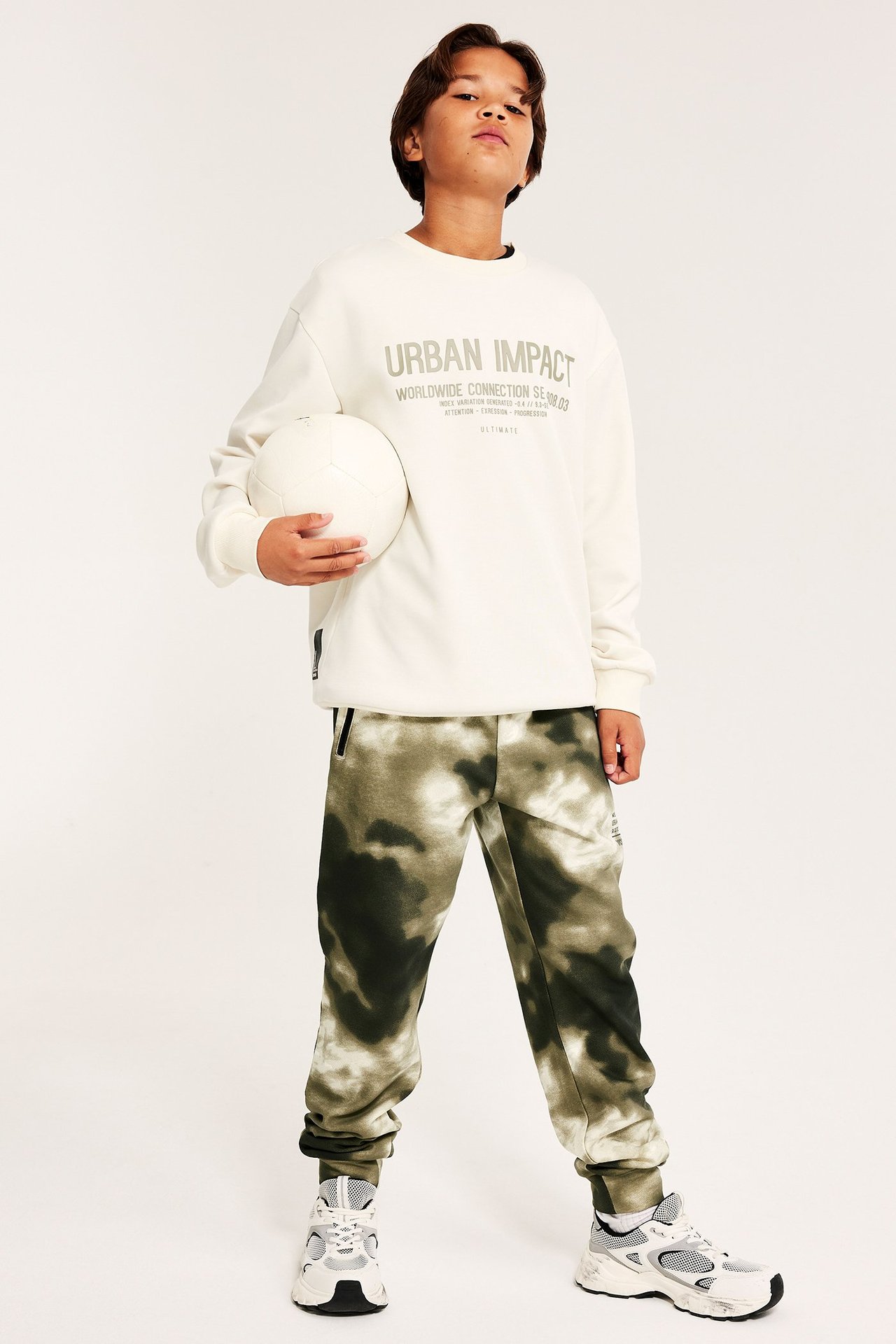 Dziecko w zielonych joggerach tie-dye, kremowej bluzie, białych sneakersach; trzyma białą piłkę.