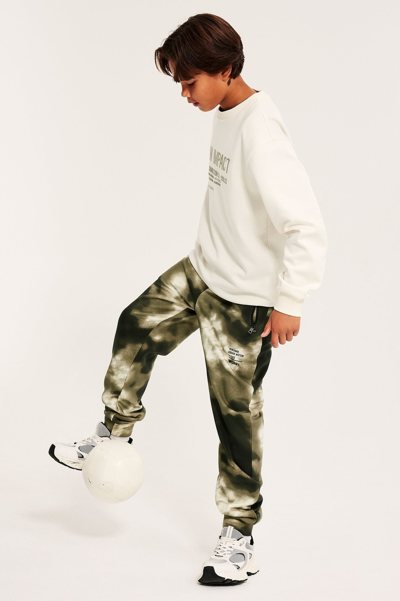 Widok z boku: Dziecko w zielonych spodniach dresowych tie-dye, bluzie ecru, białych/szarych sneakersach, kopiące piłkę.