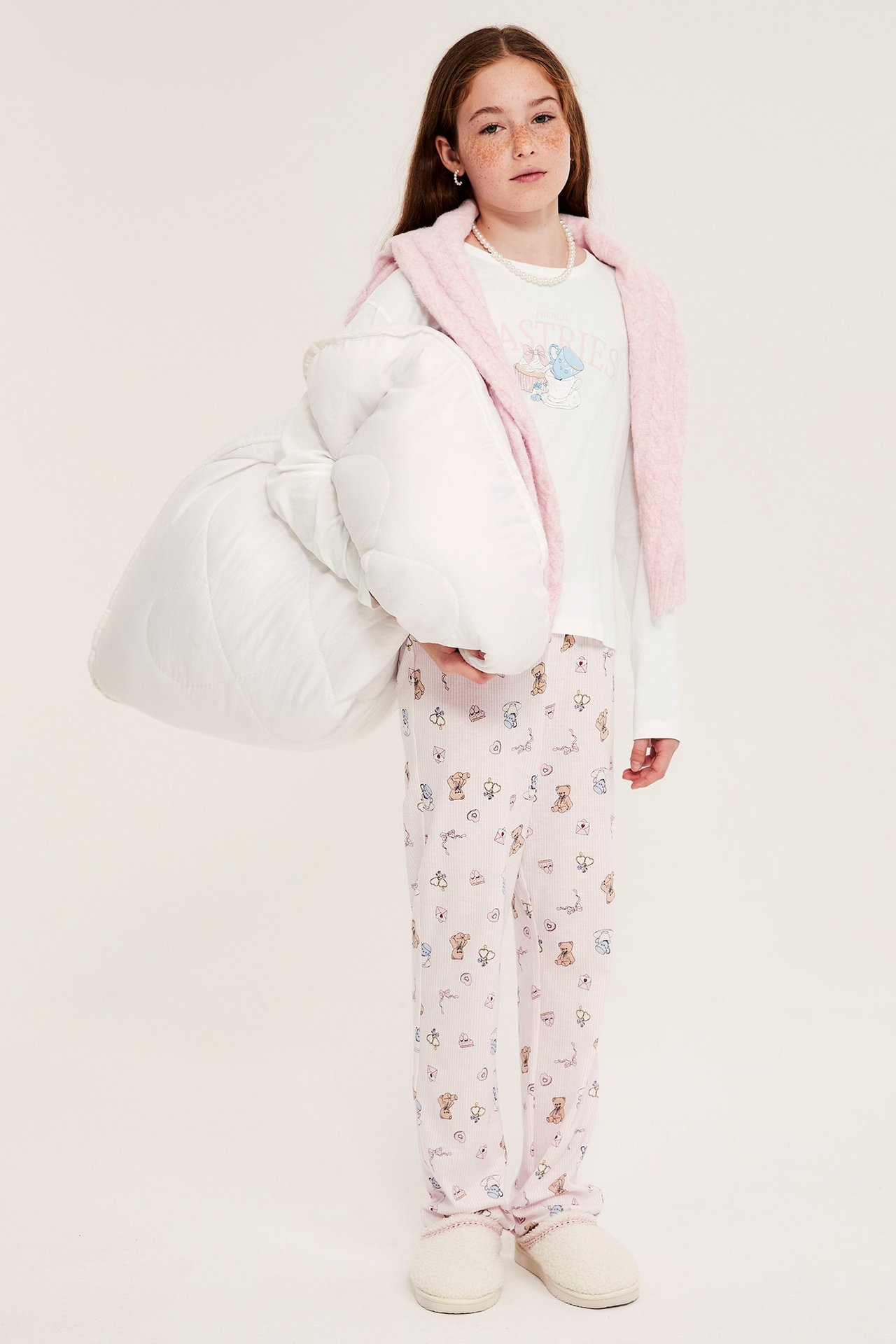 Edestä: Lapsi yllään luonnonvalkoinen pyjamas painatuksella, vaaleanpunaiset kuviolliset housut, pitäen valkoista peittoa.