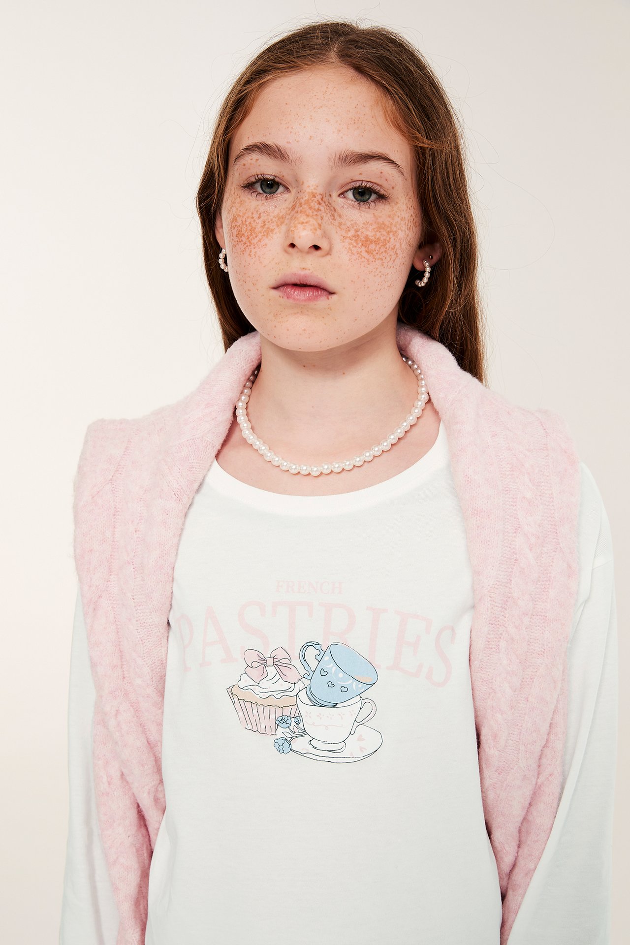 Widok z przodu: Dziecko w kremowym topie piżamowym z nadrukiem, różowym szalu i perłowej biżuterii.