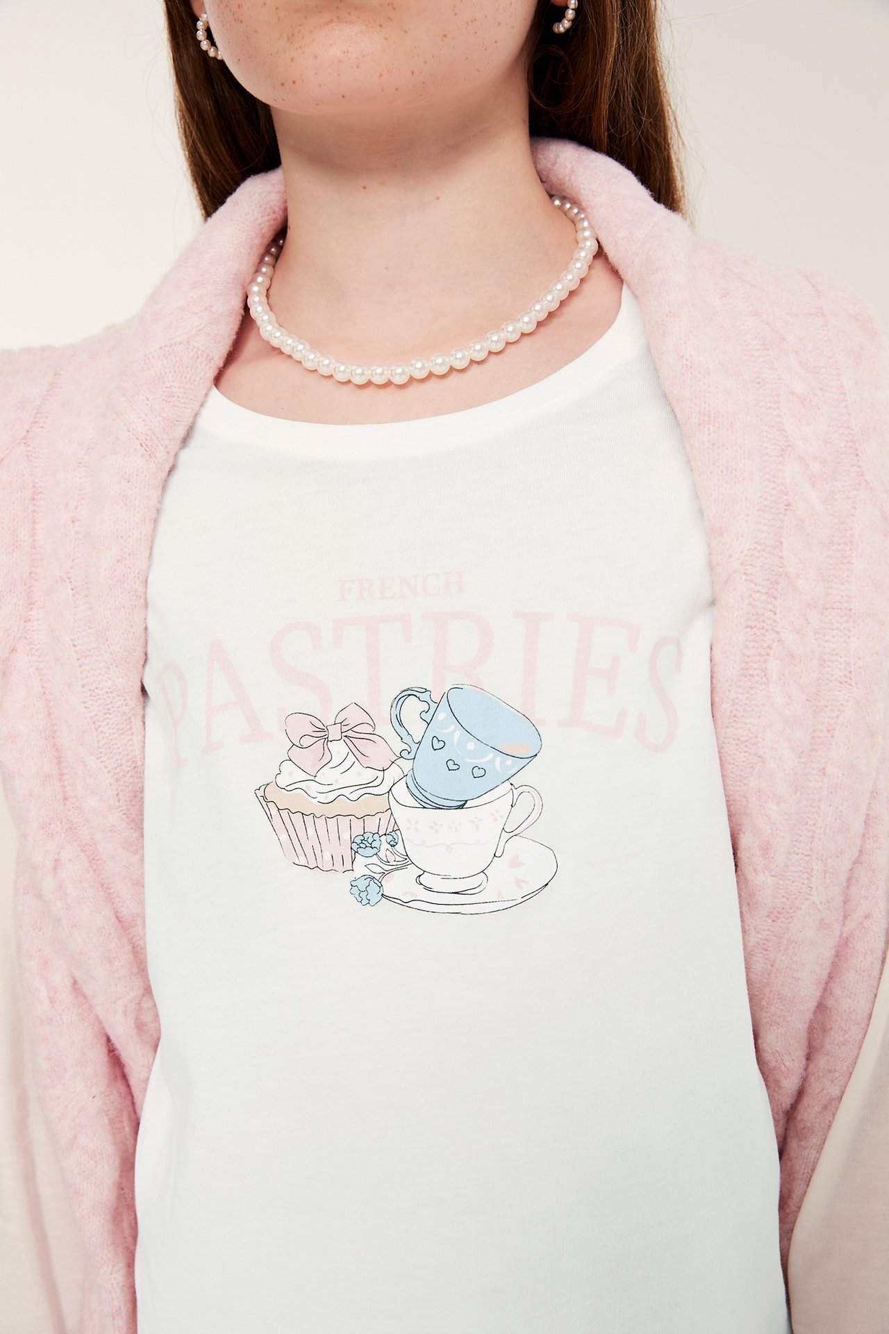 Edestä kuvattu lapsi yllään luonnonvalkoinen puuvillapyjamasyläosa "French Pastries" -printillä, vaaleanpunaisen neuletakin ja helmikaulakorun kanssa.