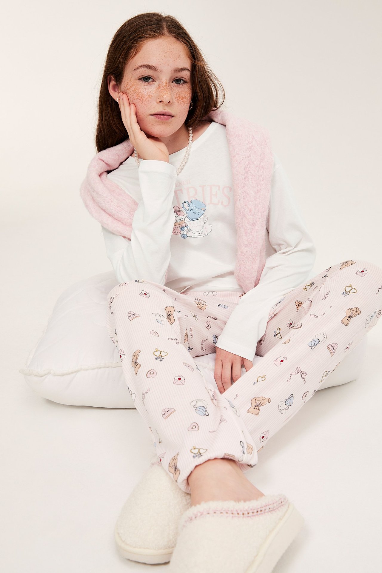 Etunäkymä: Tyttö yllään luonnonvalkoinen painettu puuvillapyjama, vaaleanpunaiset kuviolliset alaosat, pinkki huivi ja kermanväriset tohvelit.