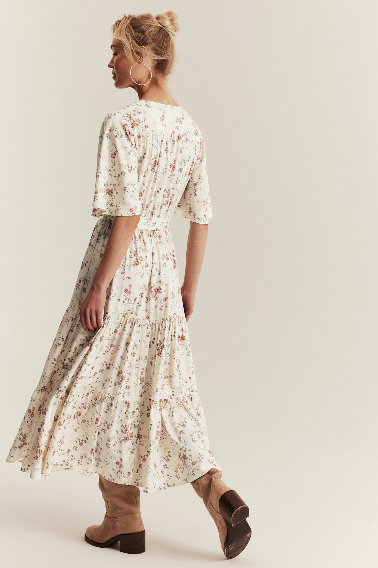 Kvinne i offwhite blomstret viskosemaxikjole med volanger og lysbrune støvler, bakre sidevisning.
