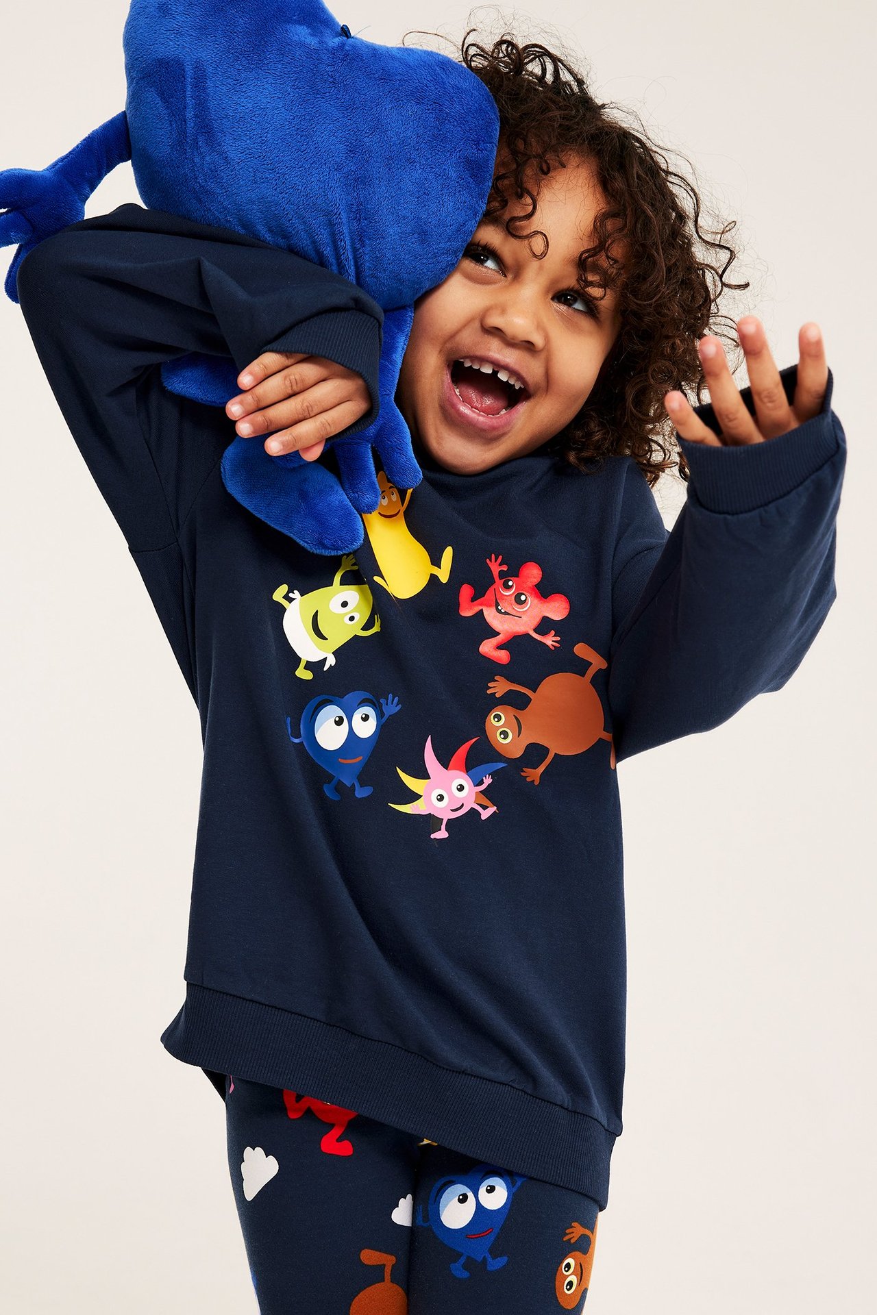 Widok z przodu: Dziecko w ciemnoniebieskiej bluzie Babblarna i pasujących legginsach, trzyma niebieską maskotkę.