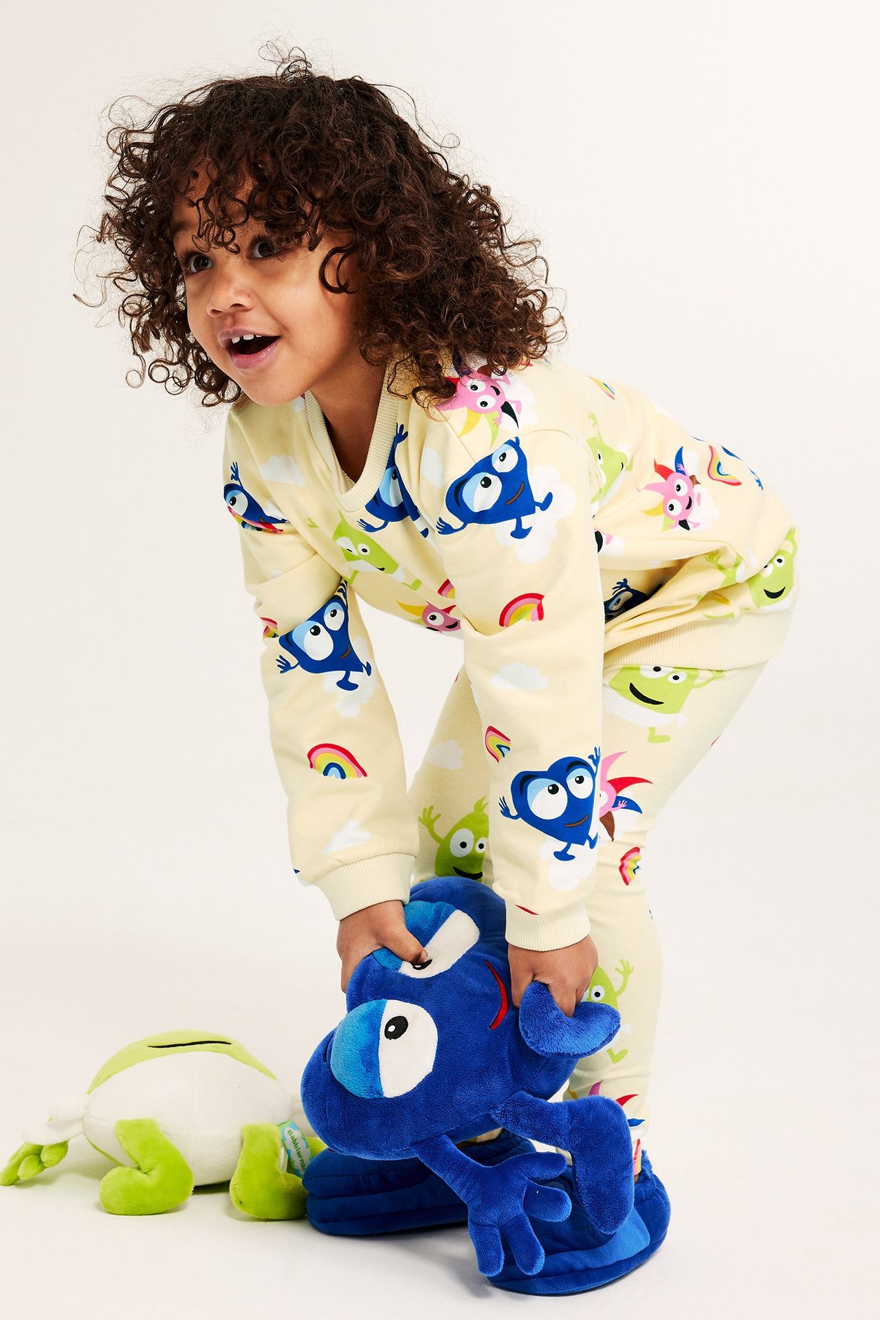 Frontvy av ett barn i en ljusbeige Babblarna-mönstrad sweatshirt och matchande byxor, hållandes en blå mjukdjur.
