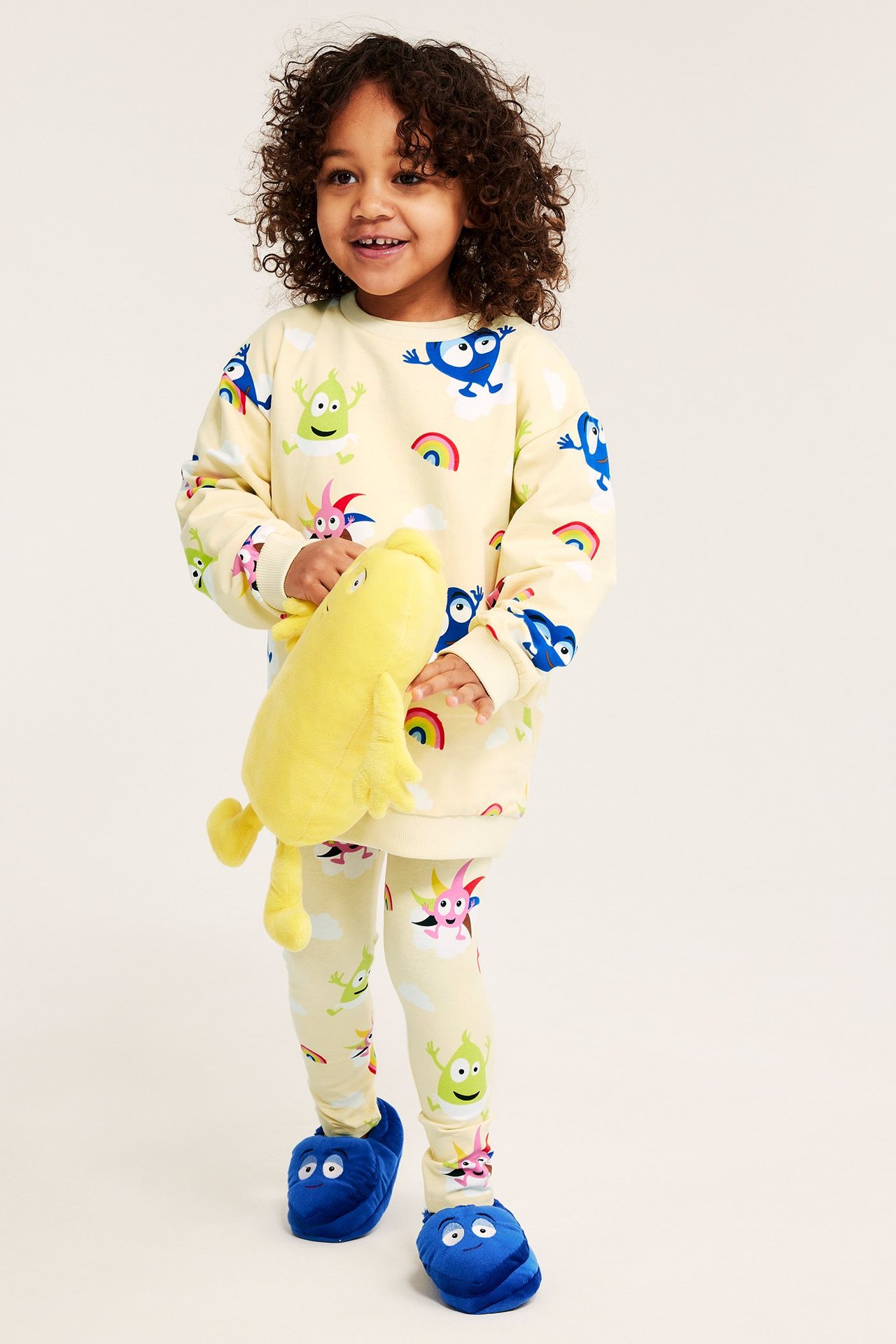 Framifrån: Kid i ljusbeige Babblarna-mönstrad sweatshirt, matchande leggings, blå tofflor, håller en gul mjukis.