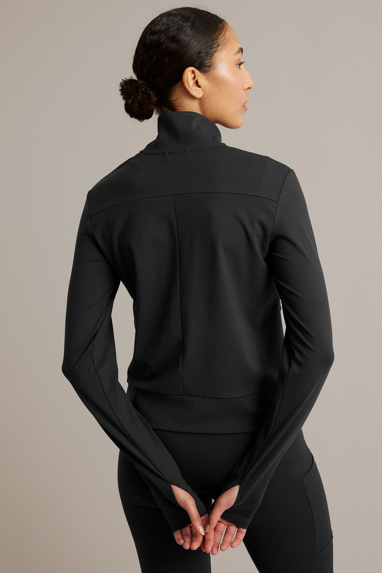 Sida-bakifrån vy av kvinna i svart träningsjacka med hög krage, tumhål och svarta tights.