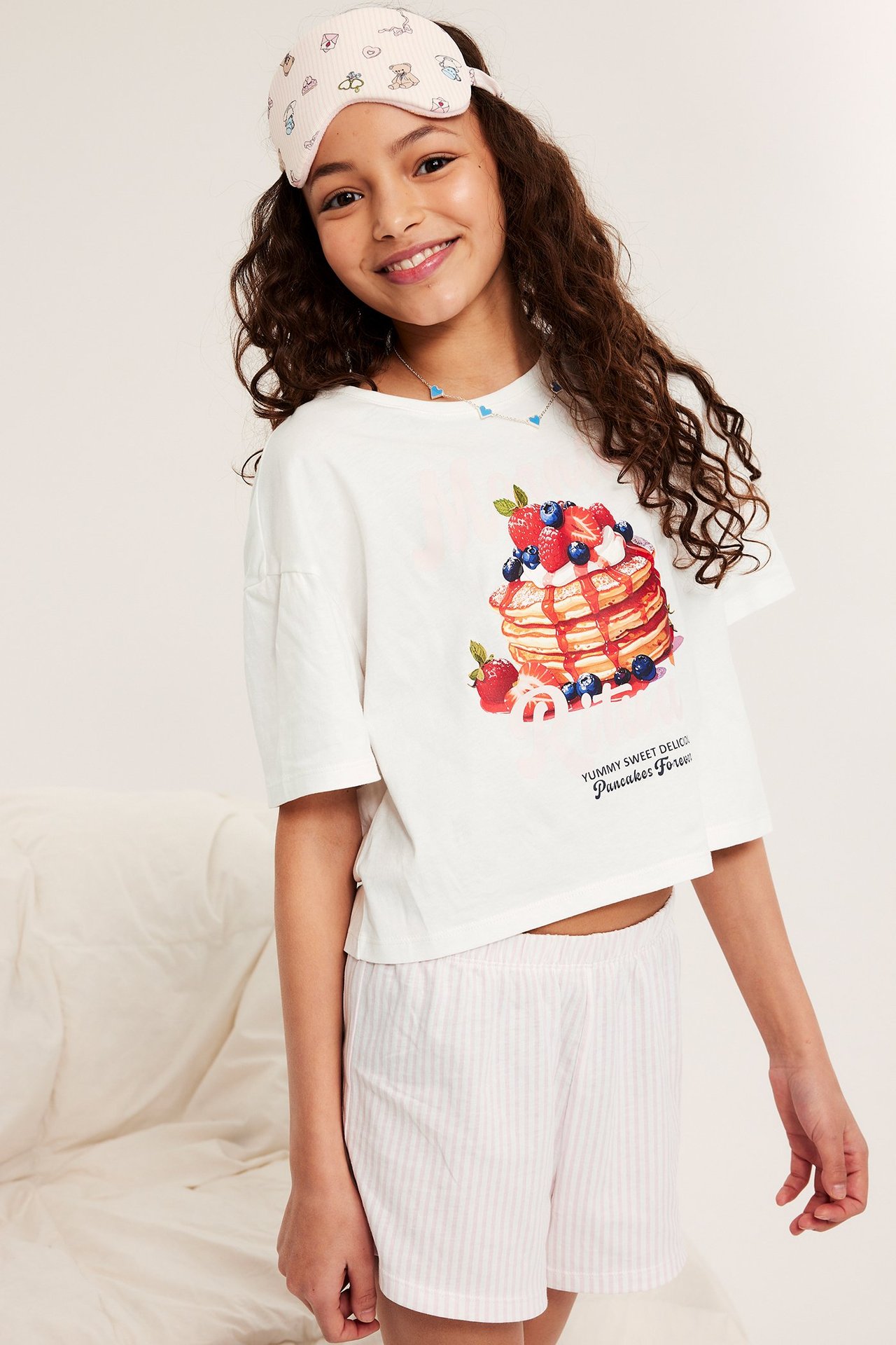 Frontbilde: Et barn i offwhite pyjamatopp i bomull, rosa stripete shorts, sovemaske og halskjede.