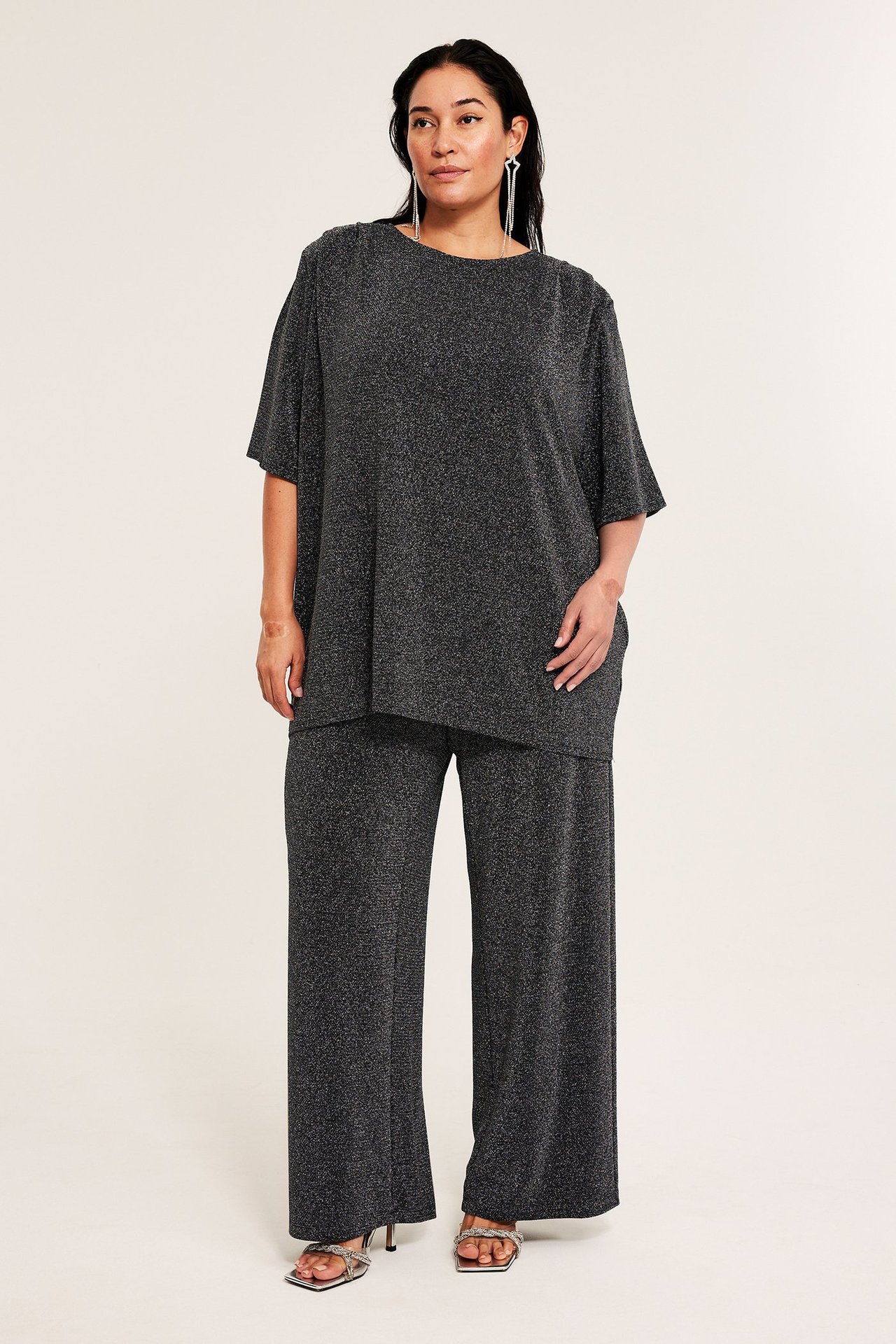 Widok z przodu: Kobieta w czarnych, błyszczących spodniach dżersejowych, pasującym topie, srebrnych kolczykach i sandałach.