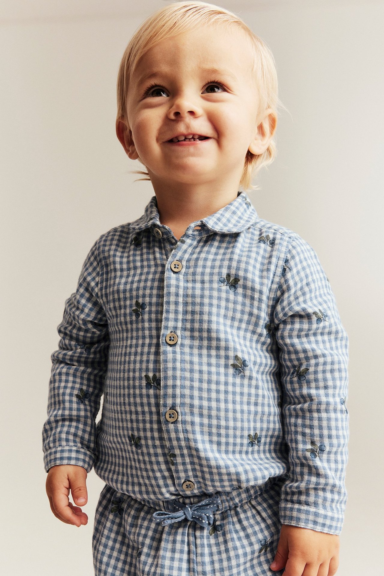 Forfra: en smilende baby i blå og hvitrutet bomullsskjortebody med blåbærtrykk og knapper.