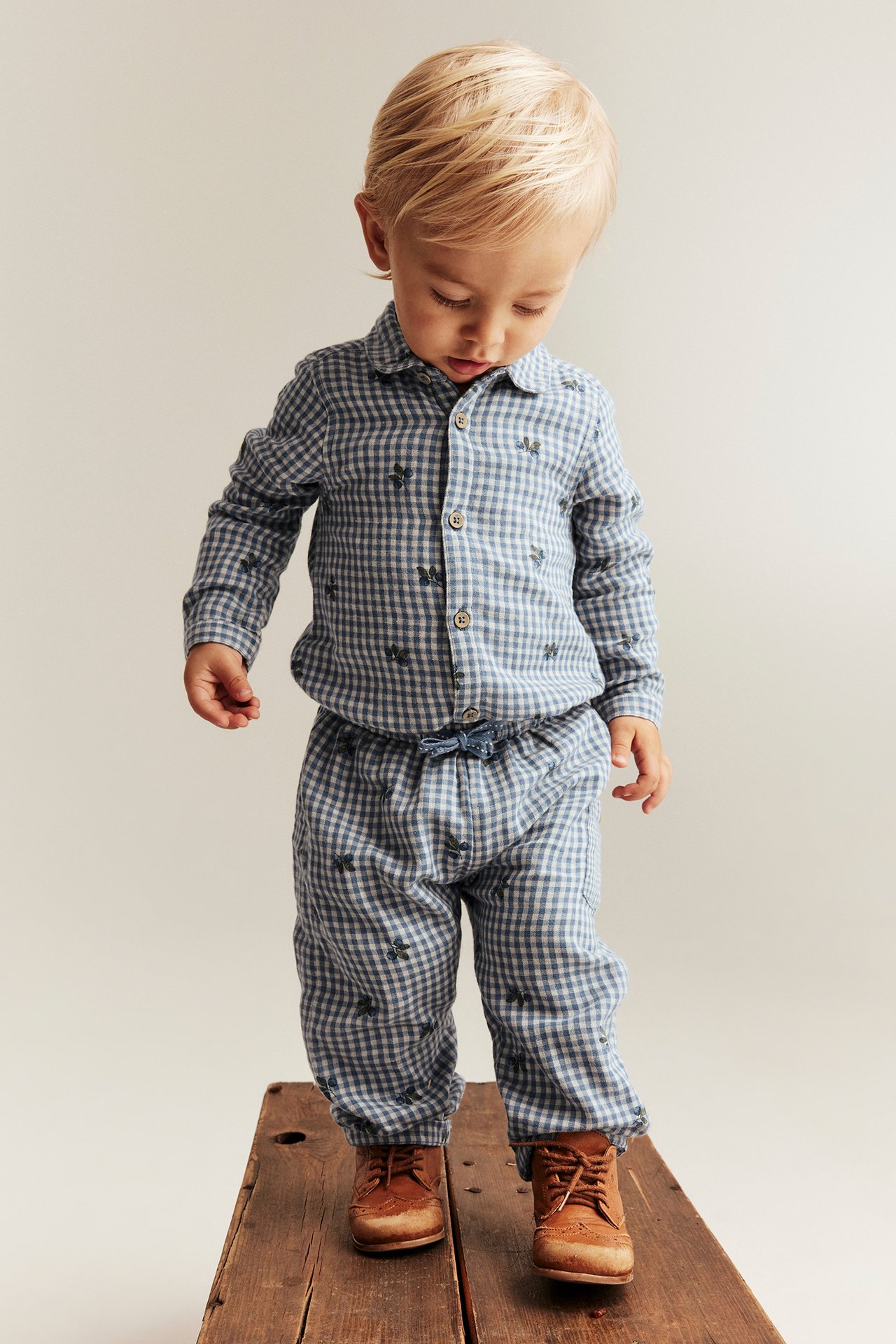 Forfra, et barn i blå rutete bomullsbody med blåbærtrykk, matchende bukser og brune snøresko.