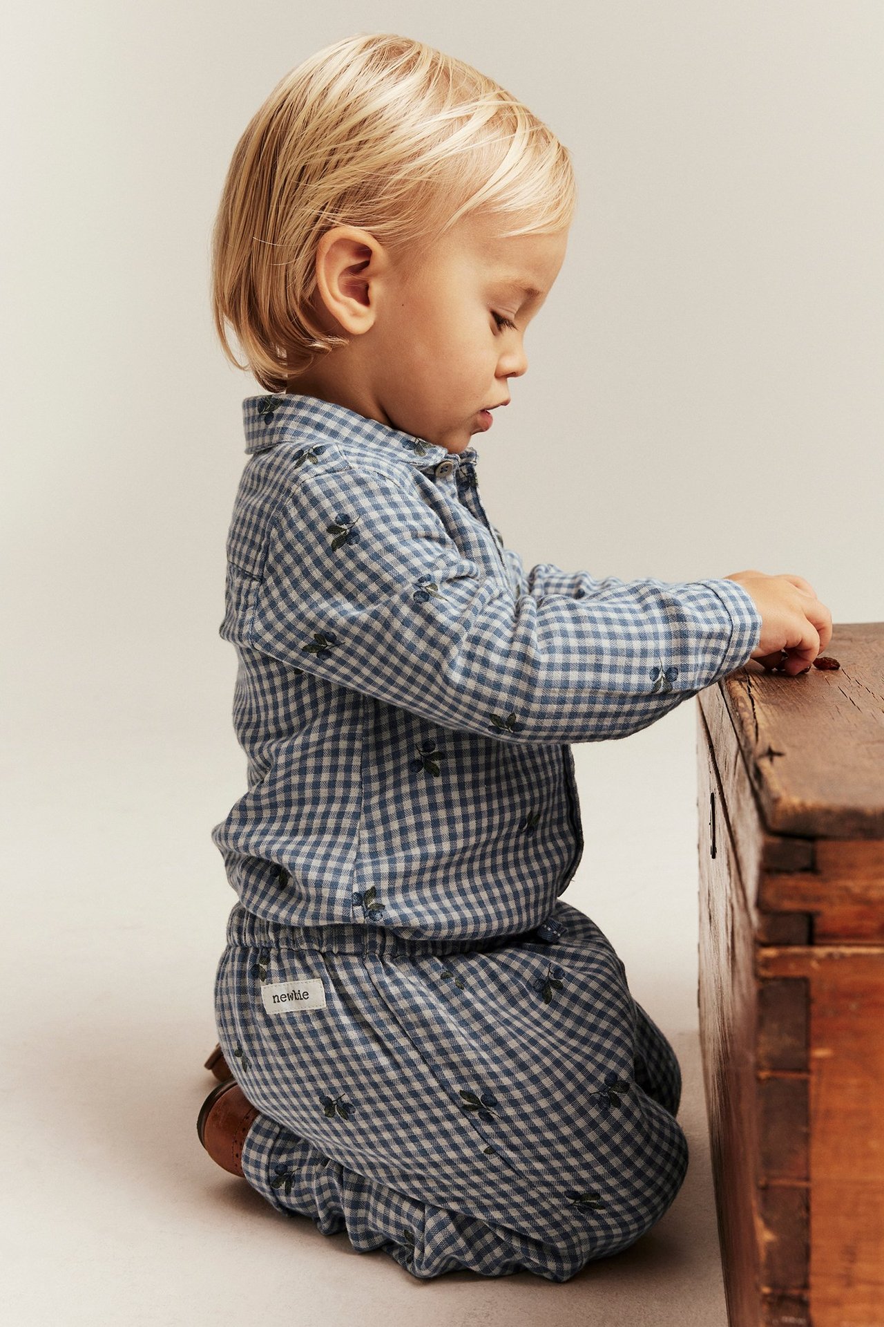 Sidebilde av et barn i blå- og hvitrutet bomulls-skjortebody med blåbærtrykk, knelende ved en trekasse.