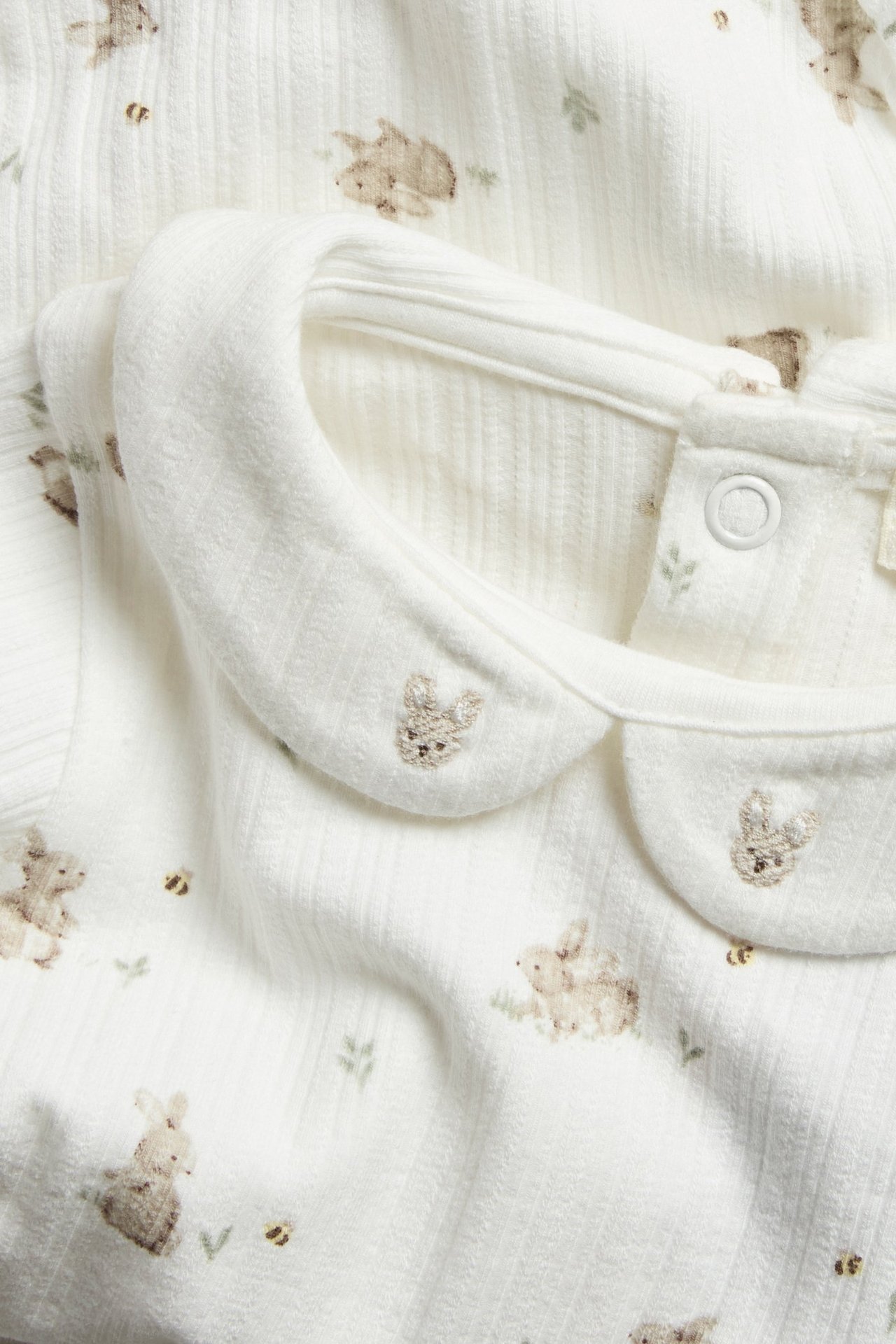 Nærbilde av en offwhite ribbet babybody med krage, broderte kaniner og trykknapp i nakken.