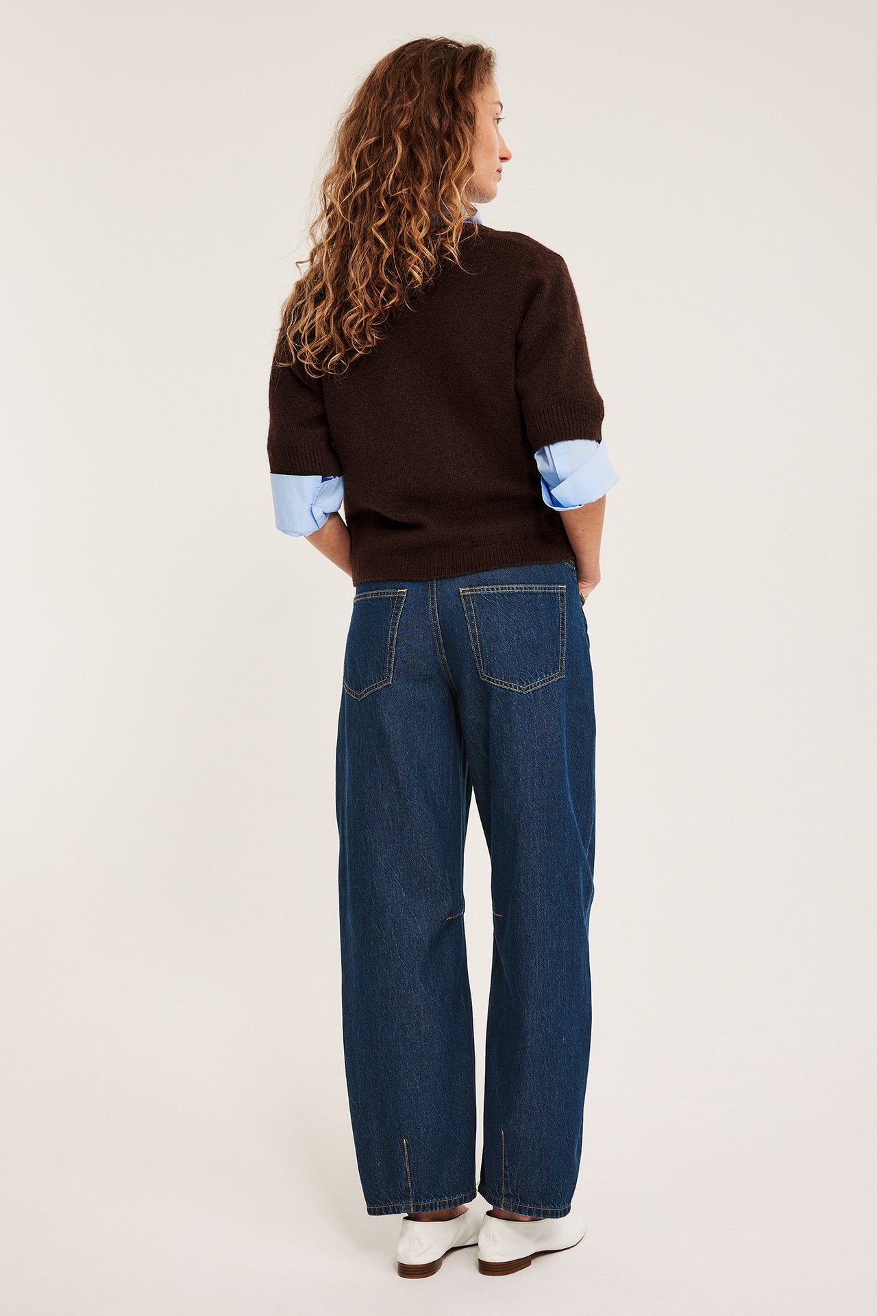 Takanäkymä: Nainen yllään tummansiniset barrel-farkut, ruskea lyhythihainen neule, vaaleansininen paita, valkoiset kengät.