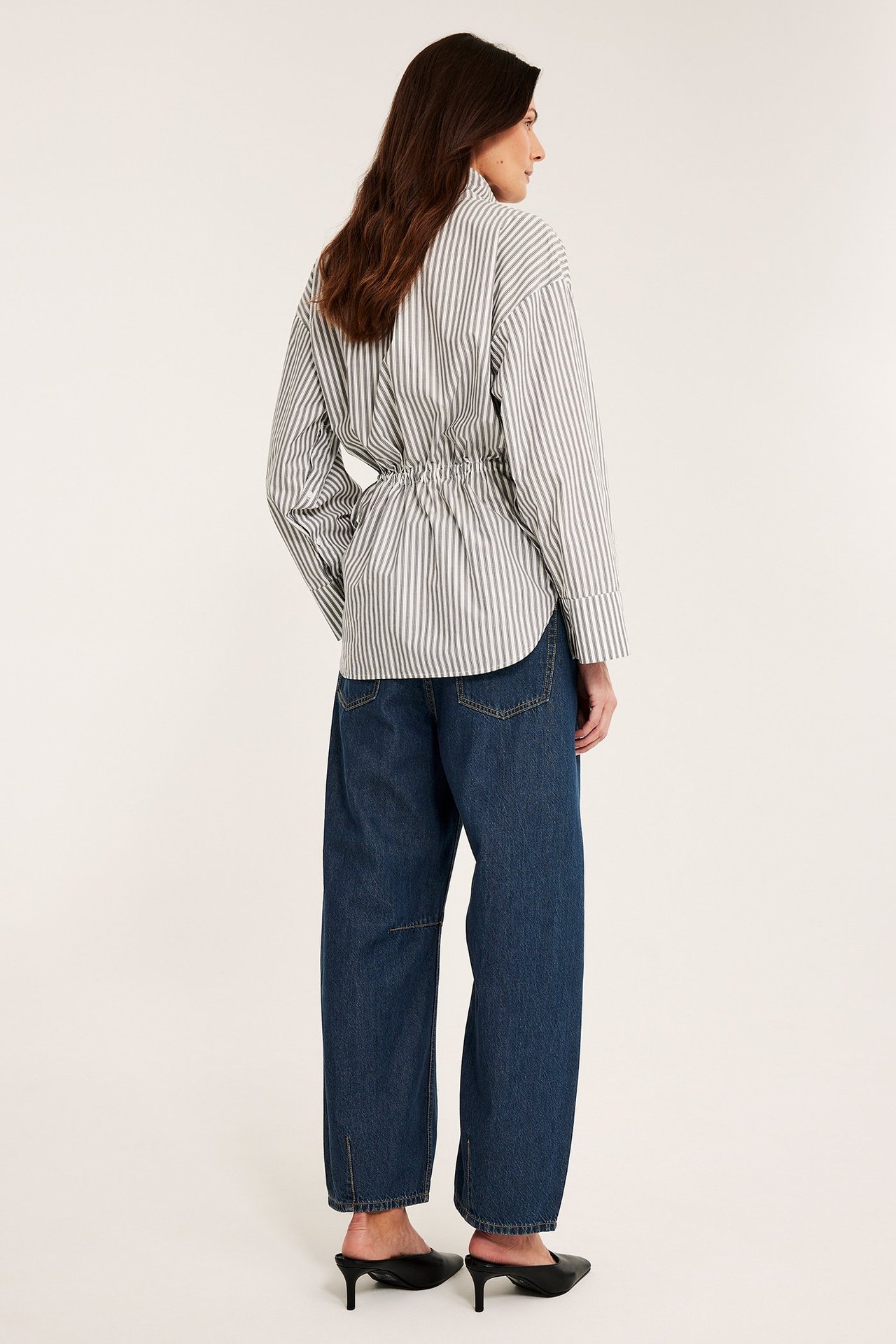 Bakside av en kvinne i mørk denim vide barrel fit-jeans, en stripete skjorte og svarte mulesko.